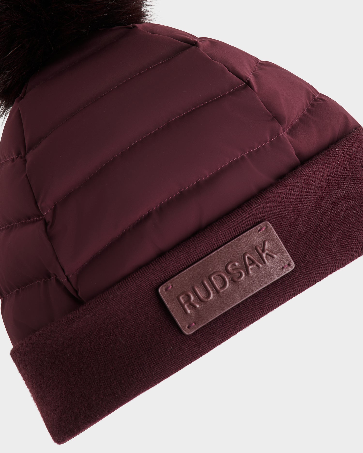 A close-up of the Rudsak GALA - 8424514 Bordeaux womens quilted down puffer hat with a folded brim, leather Rudsak patch, and a dark faux fur pom pom on top for style.