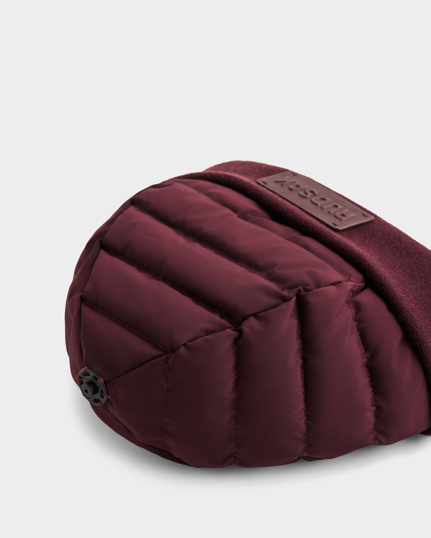 A close-up of the GALA - 8424514 Bordeaux by Rudsak, a maroon quilted bag with a soft, padded texture and black plastic fastener. The strap has a brown leather patch embossed with the Rudsak brand name. Set against a plain white background.