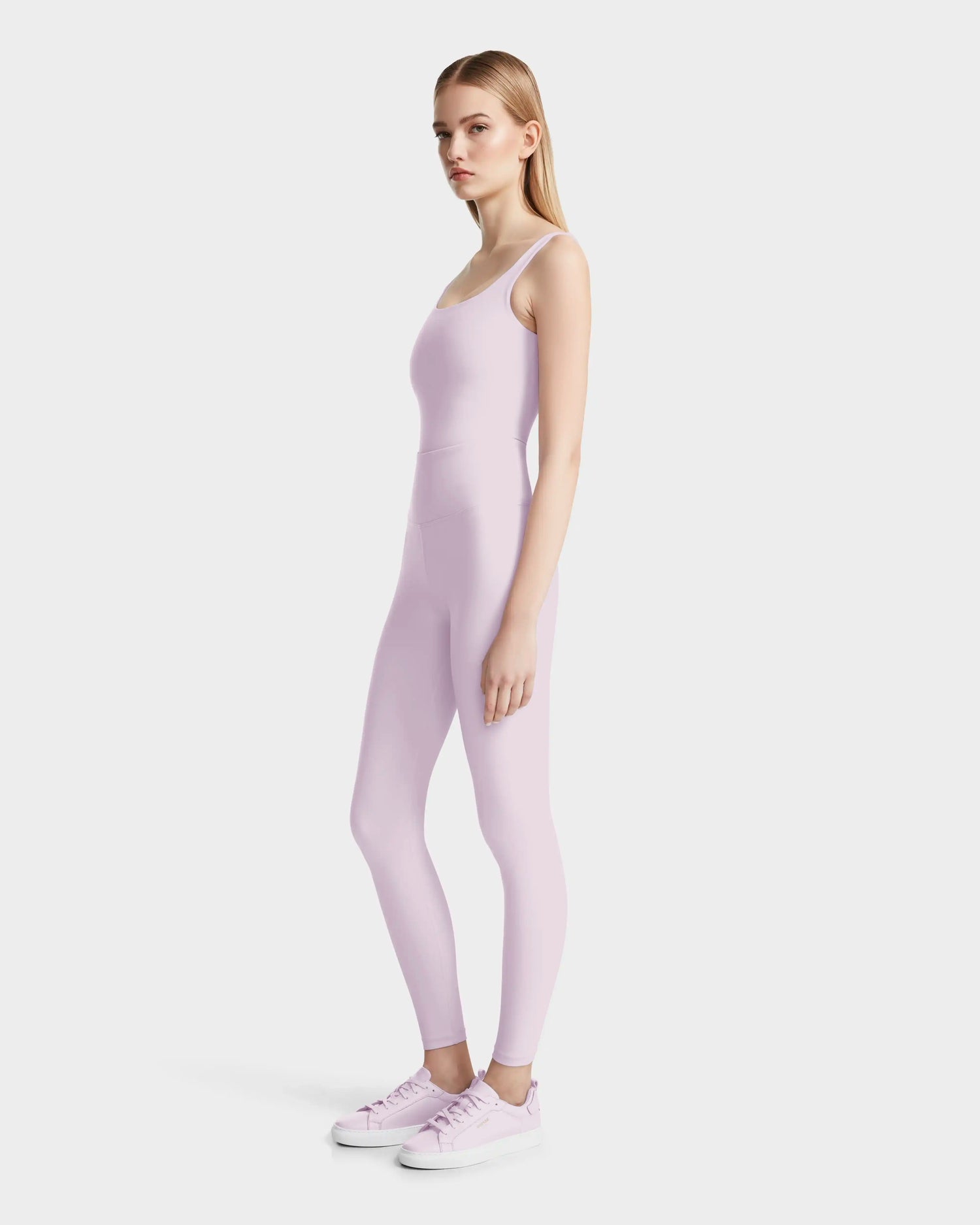 A woman stands sideways against a plain white background, wearing the BRIDGET - 8524033 Lilac Foam fitted bodysuit by Rudsak with matching sneakers.