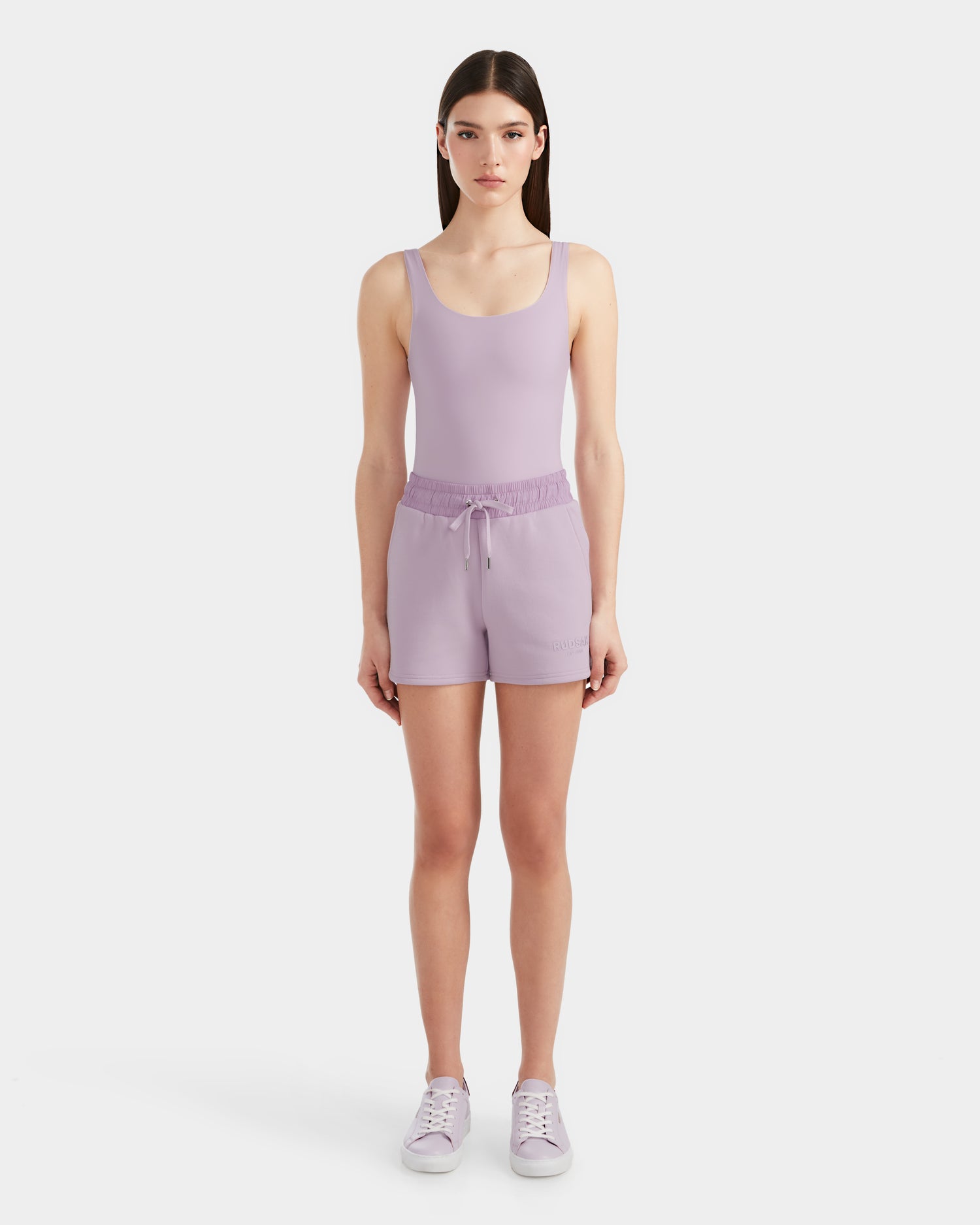 A woman faces forward in a Rudsak GENEVA - 8524070 Lilac Foam tank top, sporty drawstring shorts, and purple sneakers, standing against a plain white background.