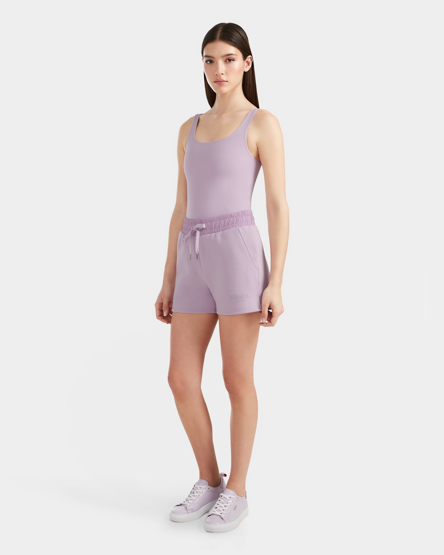A woman with long brown hair wears the Rudsak GENEVA - 8524070 Lilac Foam tank top with sporty shorts and matching sneakers, standing against a plain white background.