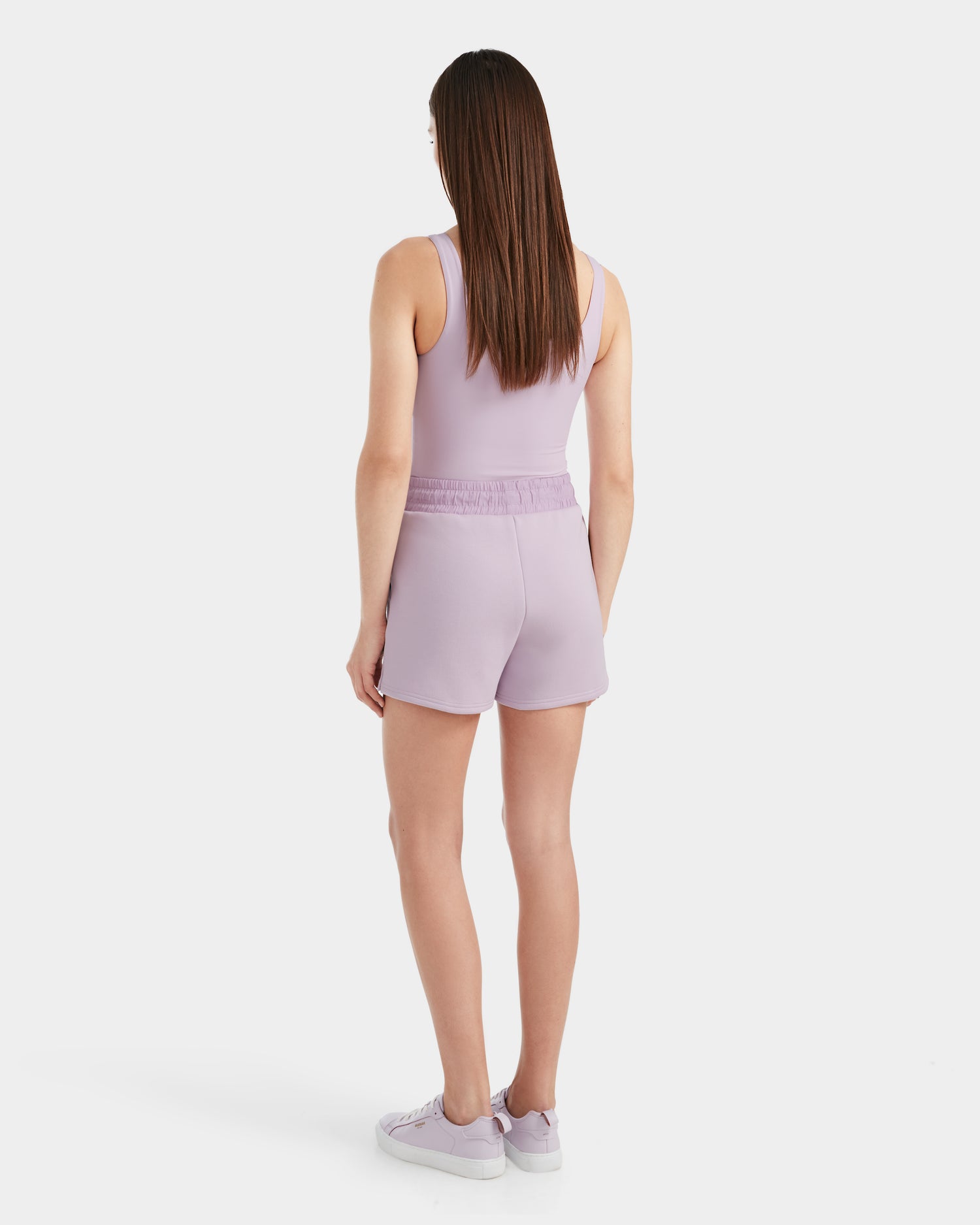 A woman with straight brown hair faces away, wearing the Rudsak GENEVA - 8524070 Lilac Foam sleeveless top, matching womens shorts, and lavender sneakers against a plain white background.