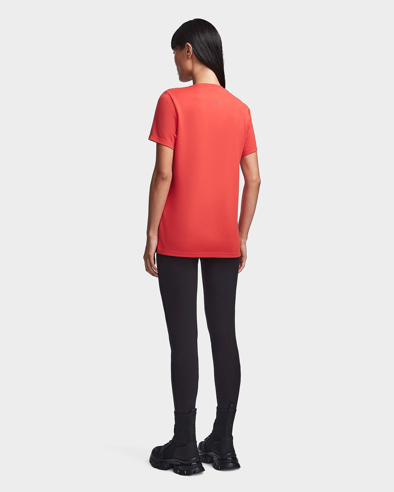 A person with long dark hair faces away, wearing a Rudsak TIFFANY - 8523048 ORANGE women’s T-shirt, black leggings, and black boots against a plain light background.