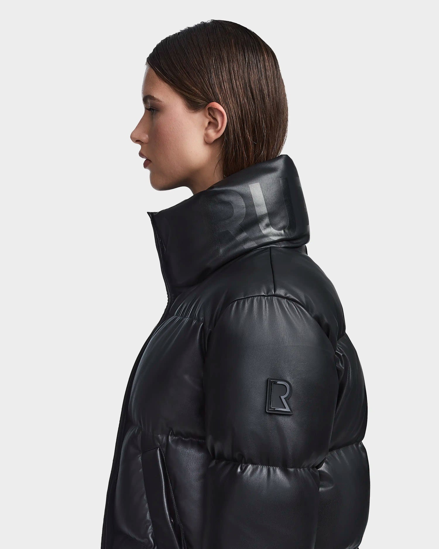 A woman with straight brown hair wears Rudsak’s GISELLE PX - L123533 Black, a shiny vegan leather puffer jacket with a high collar and embossed “R” logo, standing in profile against a plain light background—perfect for winter.