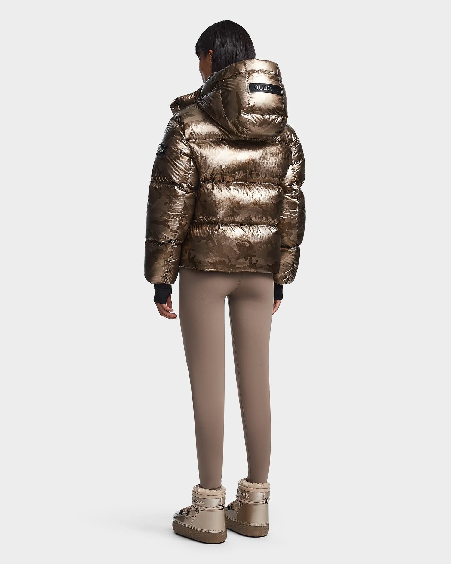 A person stands facing away, wearing the Rudsak MAVE S - 8123563 Metallic Ivory Camo women’s down puffer jacket against a plain white background.