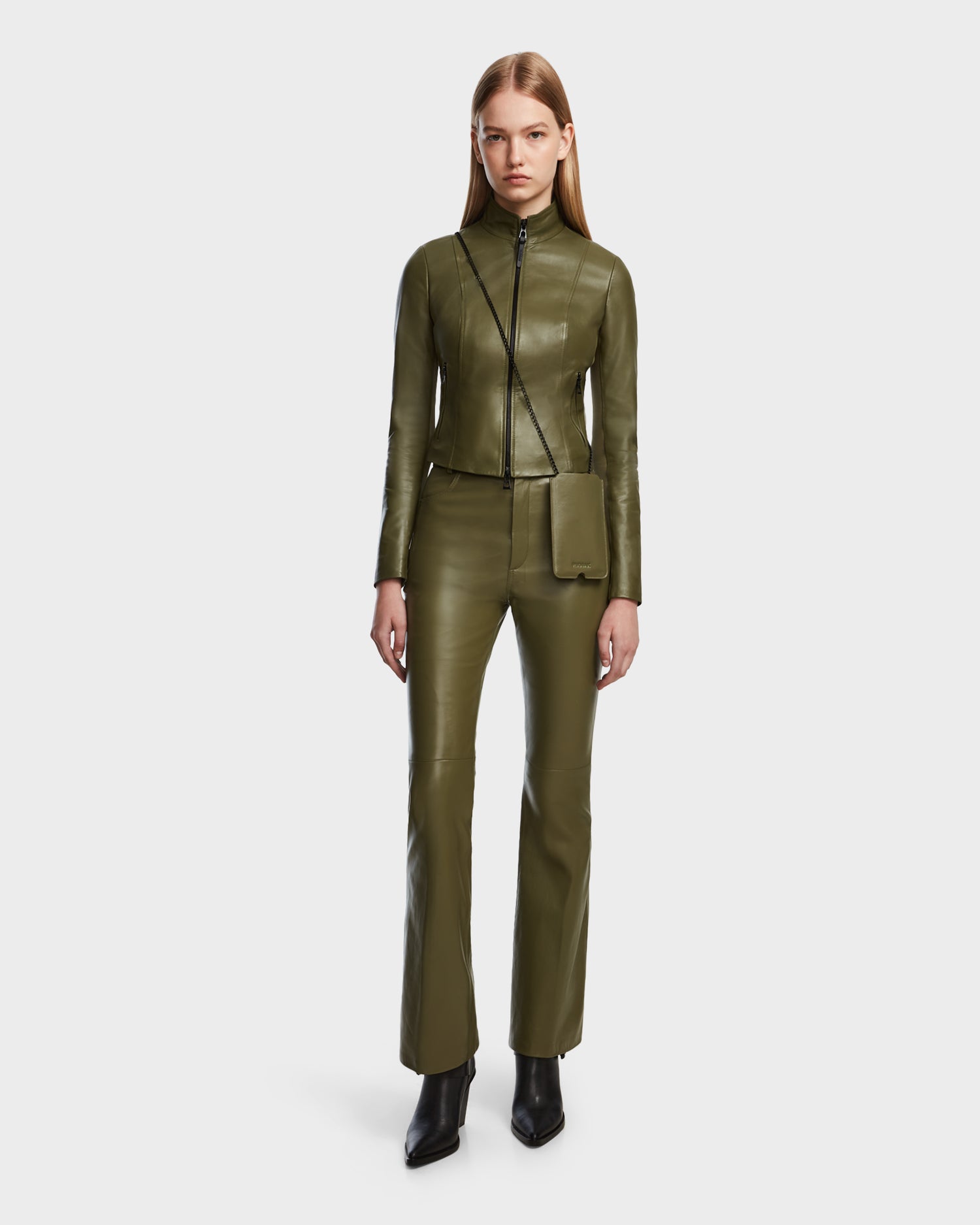 A woman with long straight hair and a neutral expression wears the Rudsak ABIGAIL - 8124560 Loden Green fitted leather jacket and matching pants, styled with a small crossbody bag and black pointed-toe boots, standing against a plain white background.