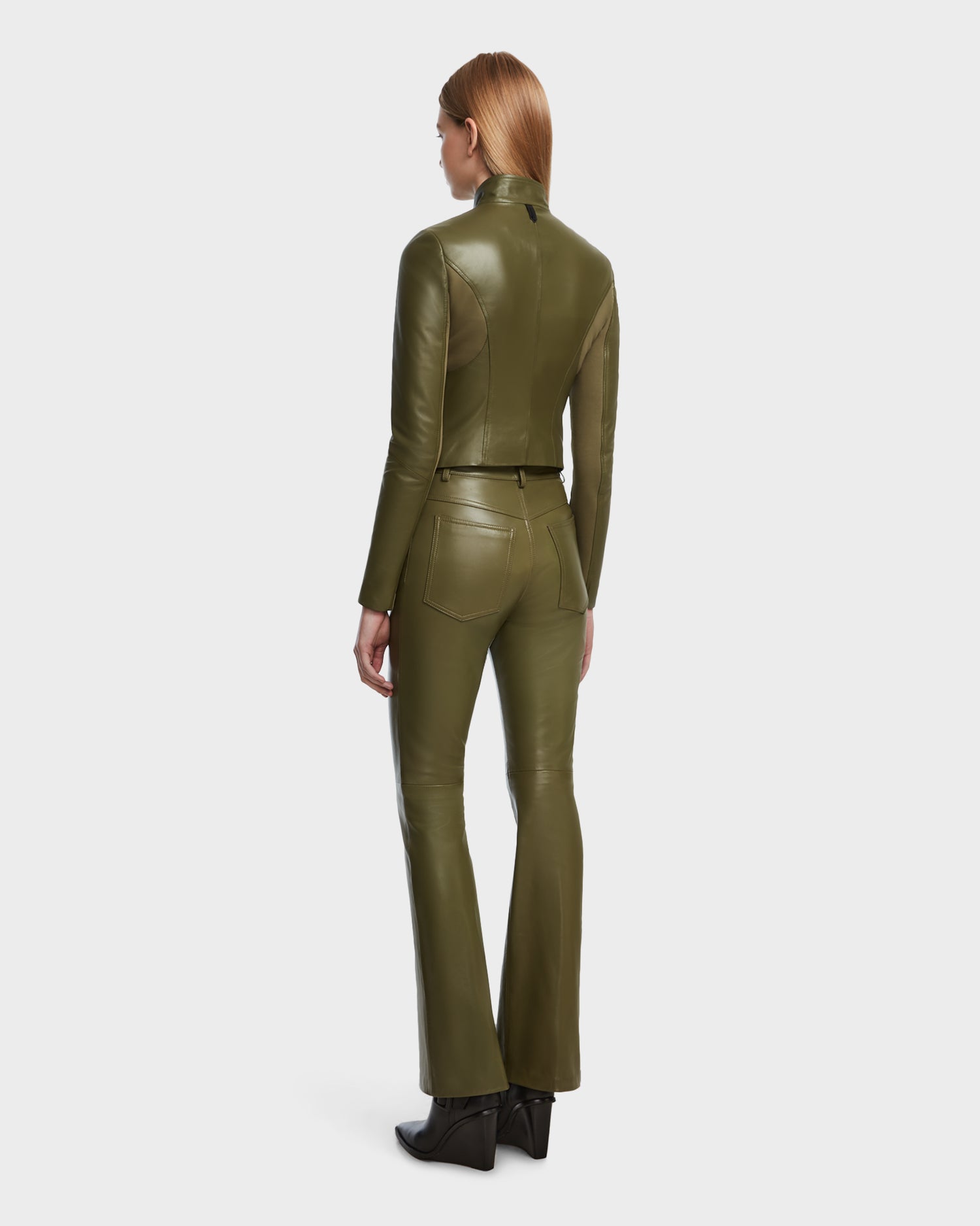 A person with straight, light brown hair stands facing away, wearing a Rudsak ABIGAIL - 8124560 Loden Green womens leather racer jacket and matching flared pants with front and back pockets, paired with black ankle boots.
