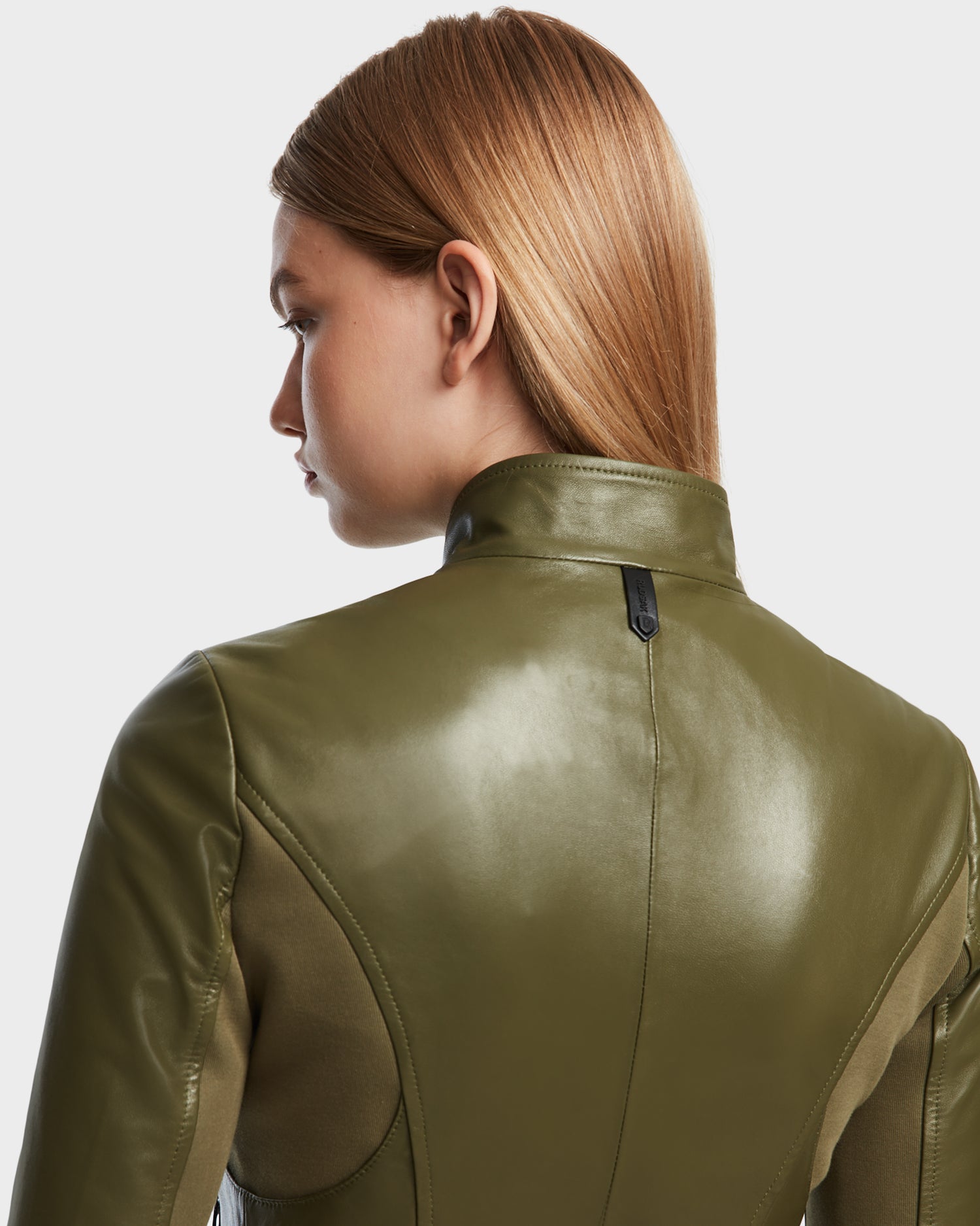 A woman with straight, light brown hair is seen from the back and side, modeling the fitted Rudsak ABIGAIL - 8124560 Loden Green leather racer jacket, which stands out against a plain, light background.