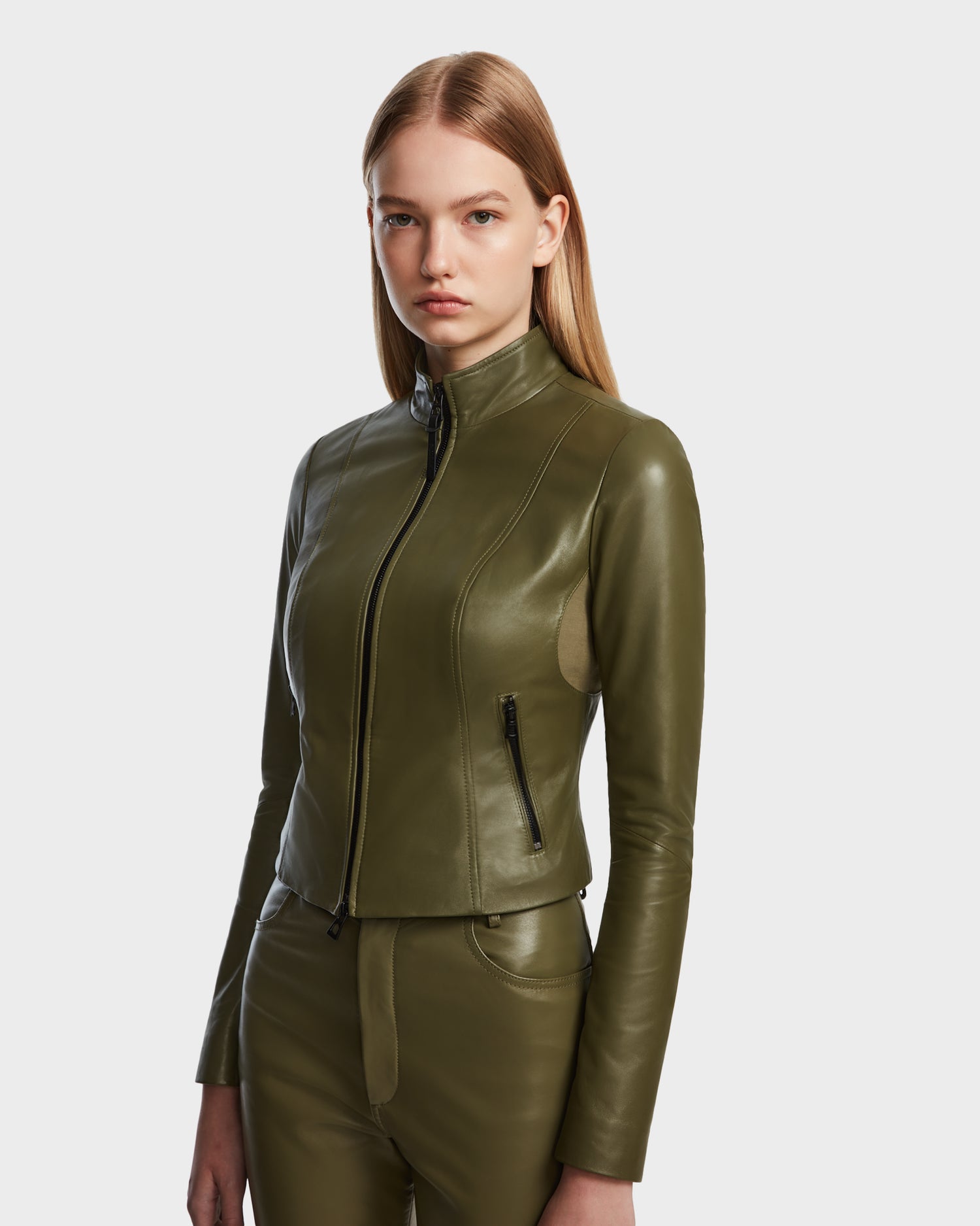 A woman with straight, light brown hair wears the Rudsak ABIGAIL - 8124560 Loden Green fitted leather jacket and matching olive green pants, standing against a plain white background.