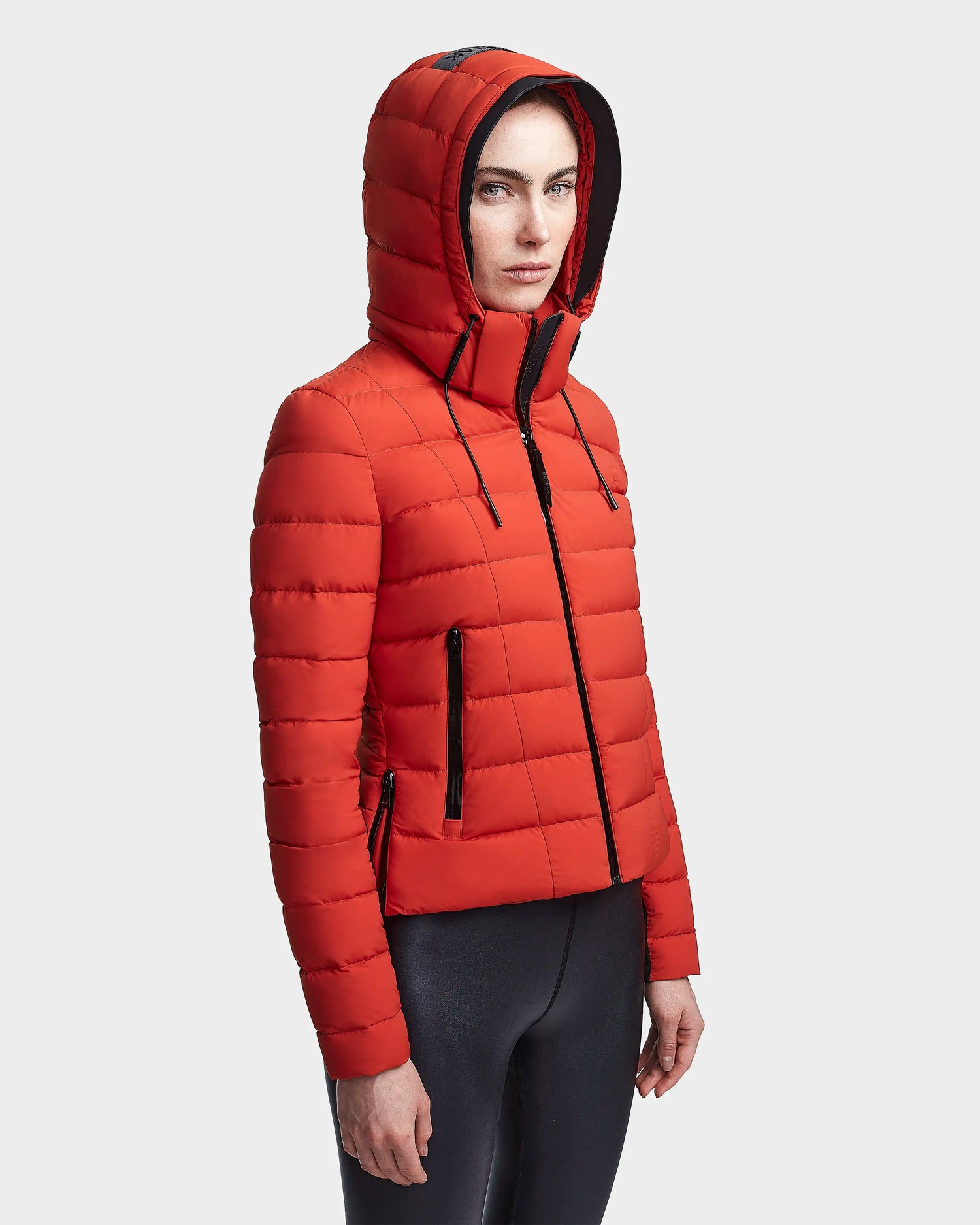 A woman wears the MELISSA - 8122551 Orange puffer jacket by Rudsak, featuring a hood and black zippers, paired with black leggings as she stands against a white background with a neutral expression.