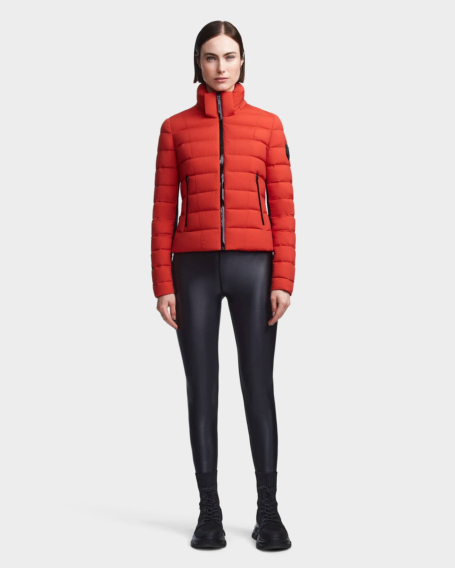 A woman faces forward in the Rudsak MELISSA - 8122551 Orange women's puffer jacket, paired with black shiny leggings and black high-top sneakers, against a plain white background.