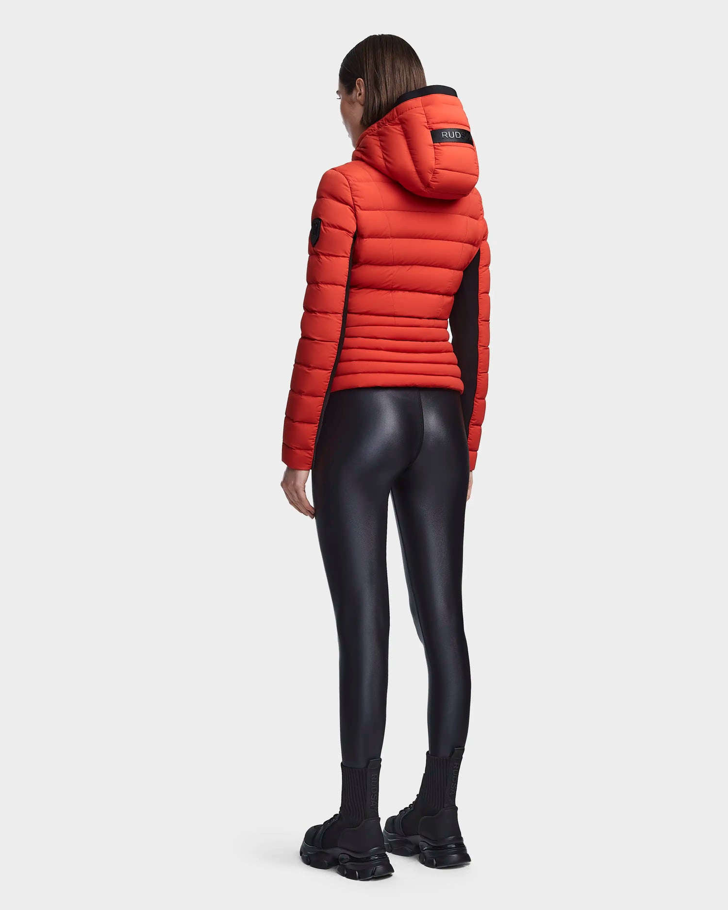 A person faces away, wearing the MELISSA - 8122551 Orange women's puffer jacket by Rudsak with a hood, black shiny leggings, black socks, and chunky shoes against a plain white background.