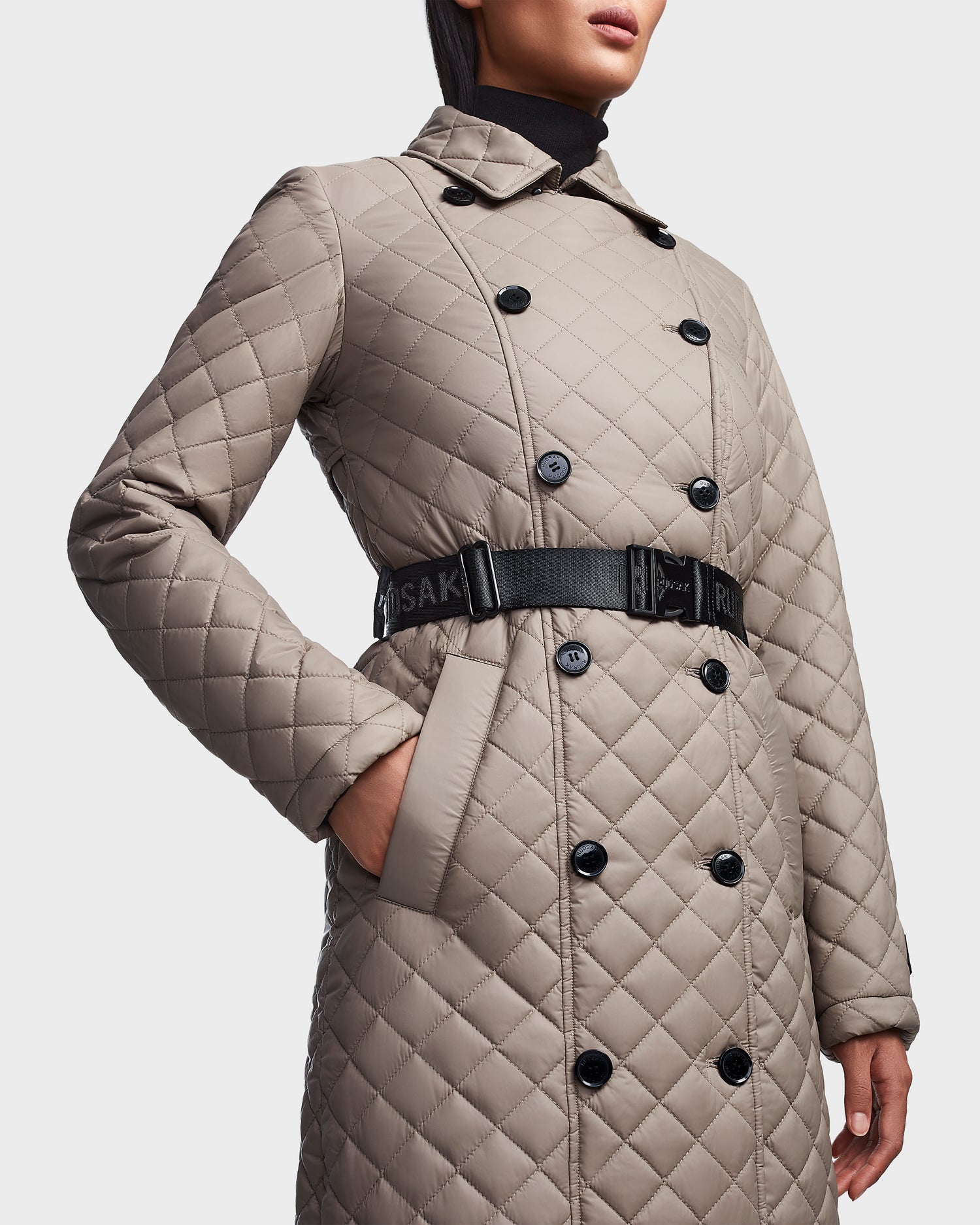 A person models the Rudsak DAISY - 8123580 Cement women's quilted maxi trench coat with black buttons, a black belt, and sustainable Sorona insulation, standing with one hand in their pocket against a plain background.