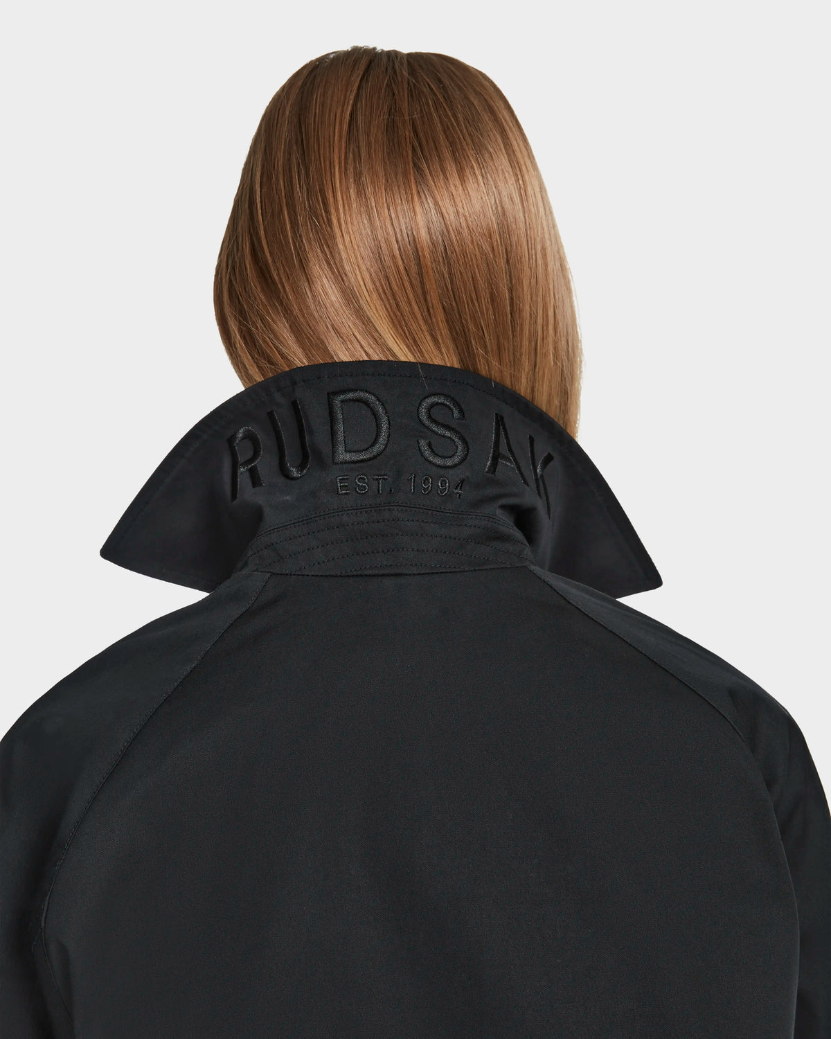A person with straight light brown hair is seen from behind wearing the Rudsak MIRELLE - 8124055 Black jacket with a tailored fit and turned-up collar featuring RUDSAK EST. 1999 embroidery, set against a plain white background.