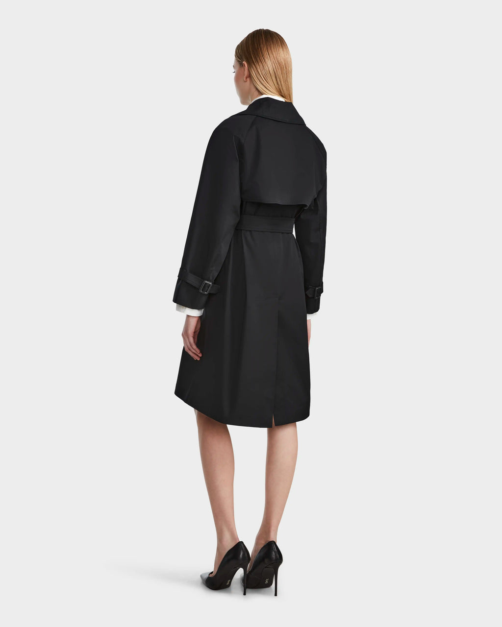 A woman with straight, light brown hair models the Rudsak MIRELLE - 8124055 Black women’s classic trench with a belted waist and black high heels, posing with her back to the camera against a plain light background.