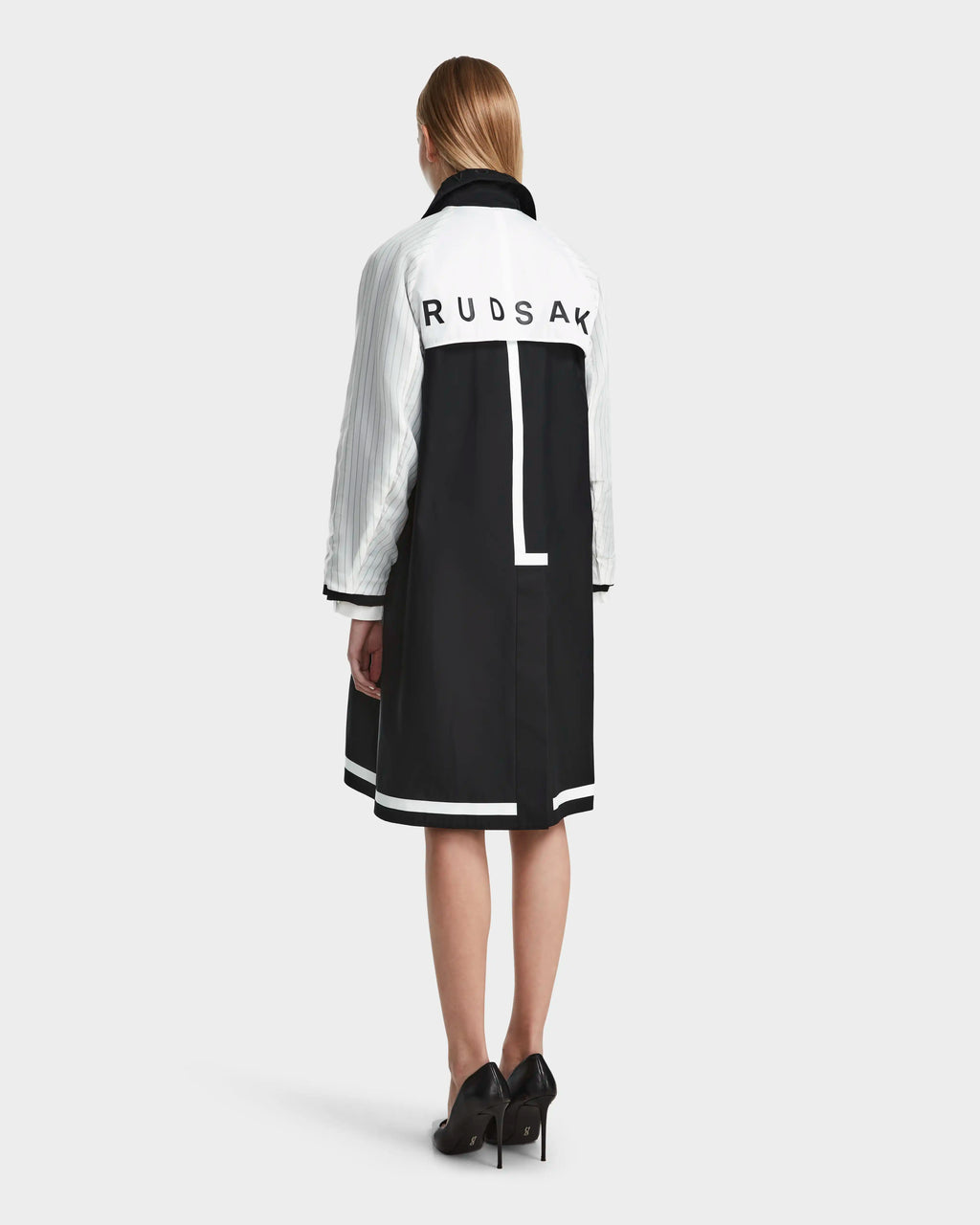A woman with straight, shoulder-length hair and black heels faces away, wearing the Rudsak MIRELLE - 8124055 Black coat featuring a tailored silhouette and logo on the back. The background is plain light gray.