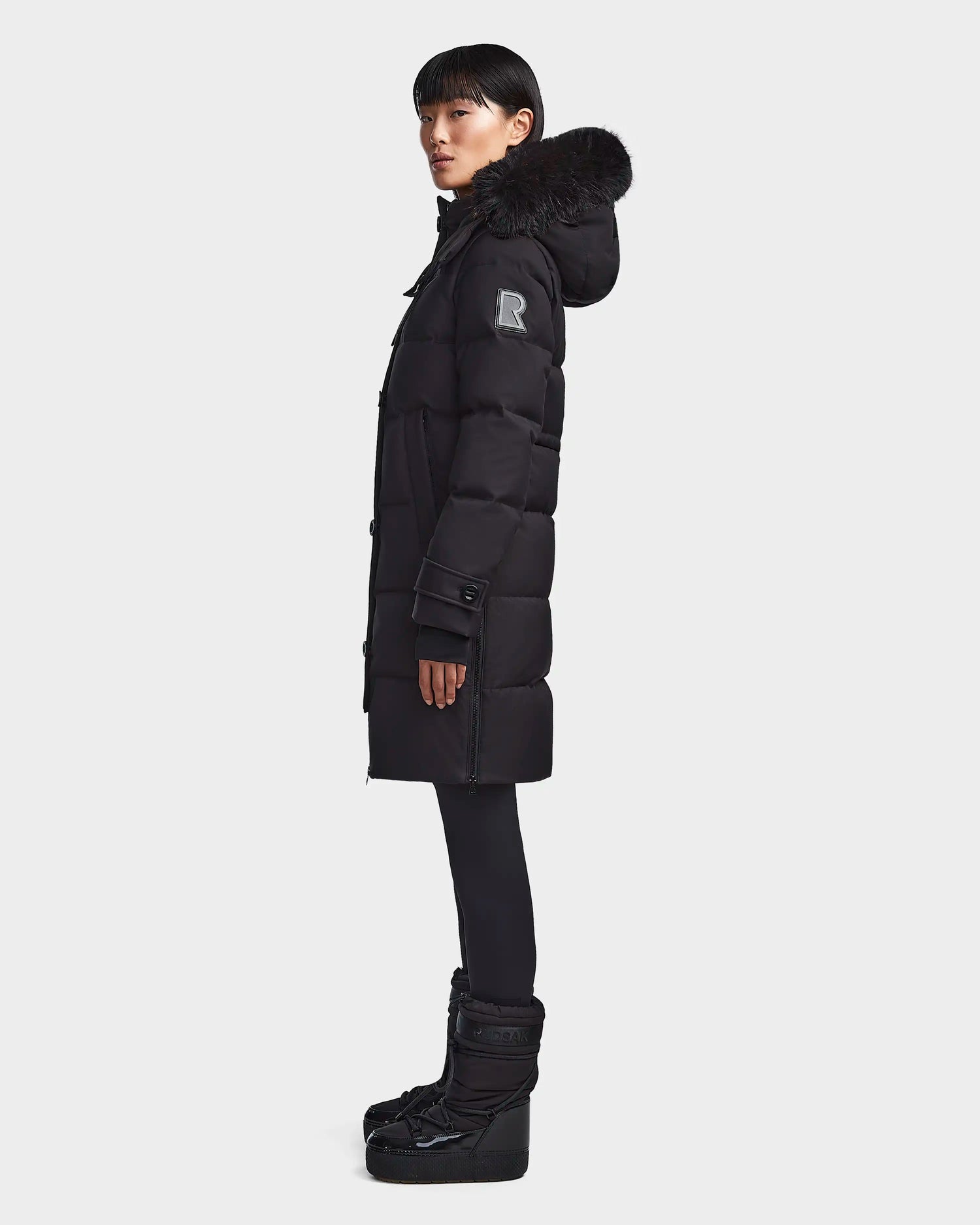 A person stands in profile against a plain light background wearing the Rudsak MEYA M - L123567 Black womens down puffer with a fur-trimmed hood, paired with black leggings and black winter boots.