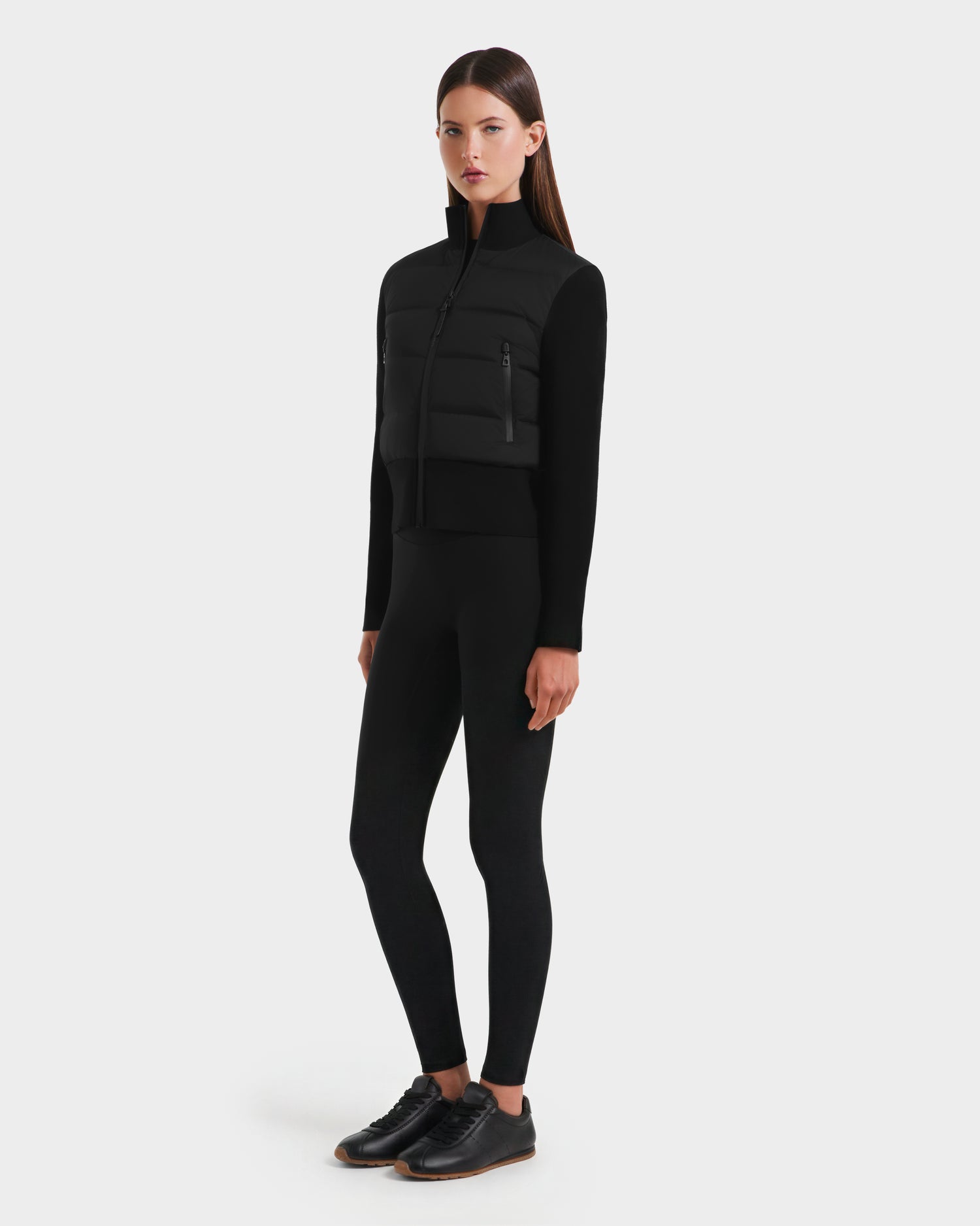 Against a plain light gray background, a woman models the Rudsak CLAREMONT - 8125502 Black women's short down jacket, paired with a black long-sleeve top, leggings, and shoes. She stands slightly turned, arms relaxed at her sides.