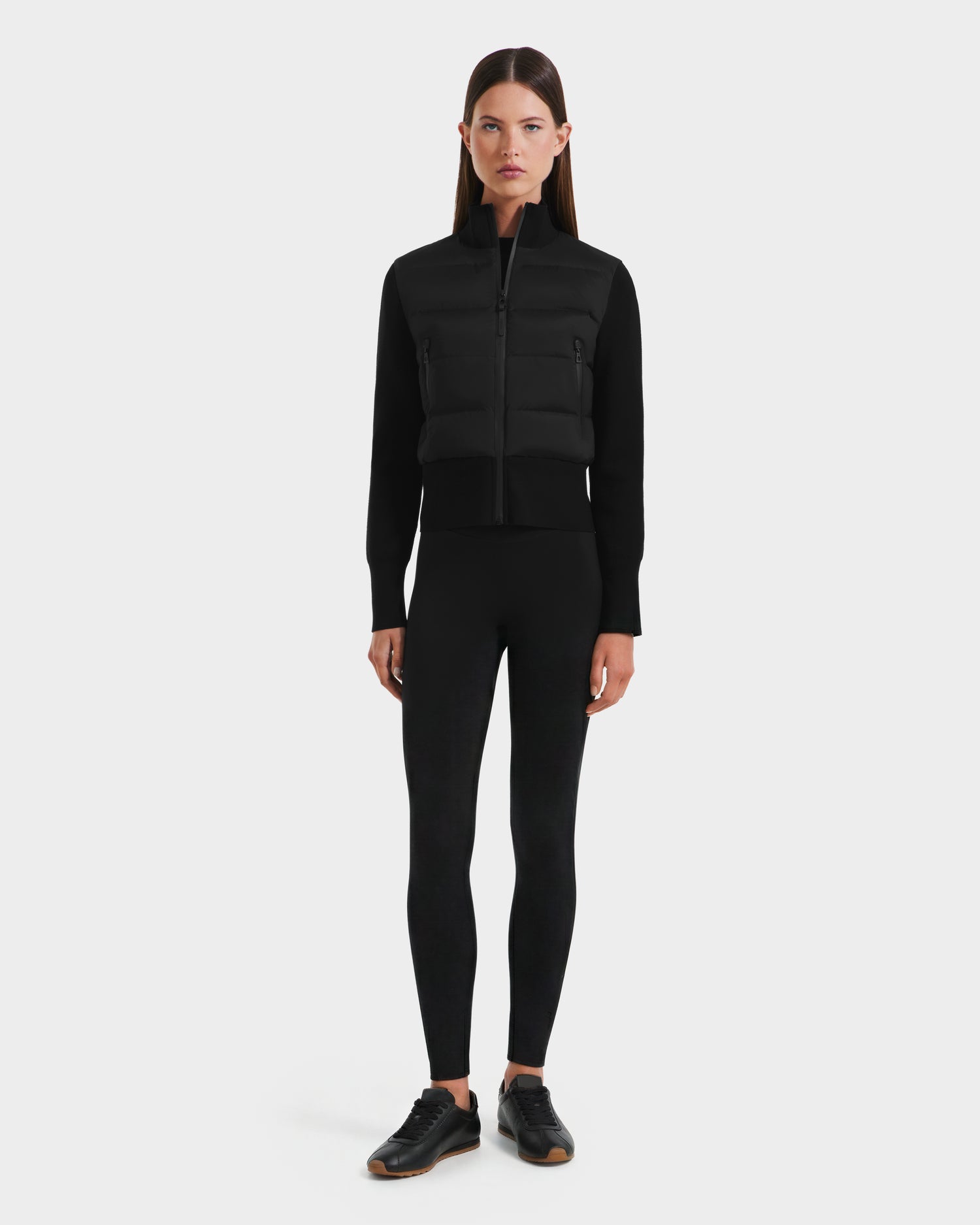 A woman faces forward wearing Rudsak's CLAREMONT - 8125502 Black hybrid down jacket, delivering lightweight warmth, paired with black leggings and sneakers against a plain light gray background.
