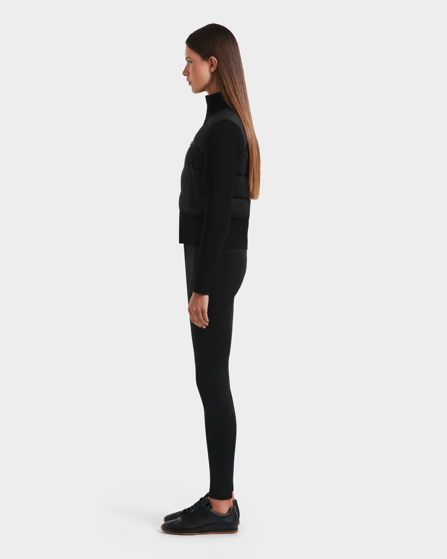 A woman in profile stands against a white background, wearing the Rudsak CLAREMONT - 8125502 Black hybrid down jacket for lightweight warmth, paired with a black turtleneck, leggings, and sneakers.