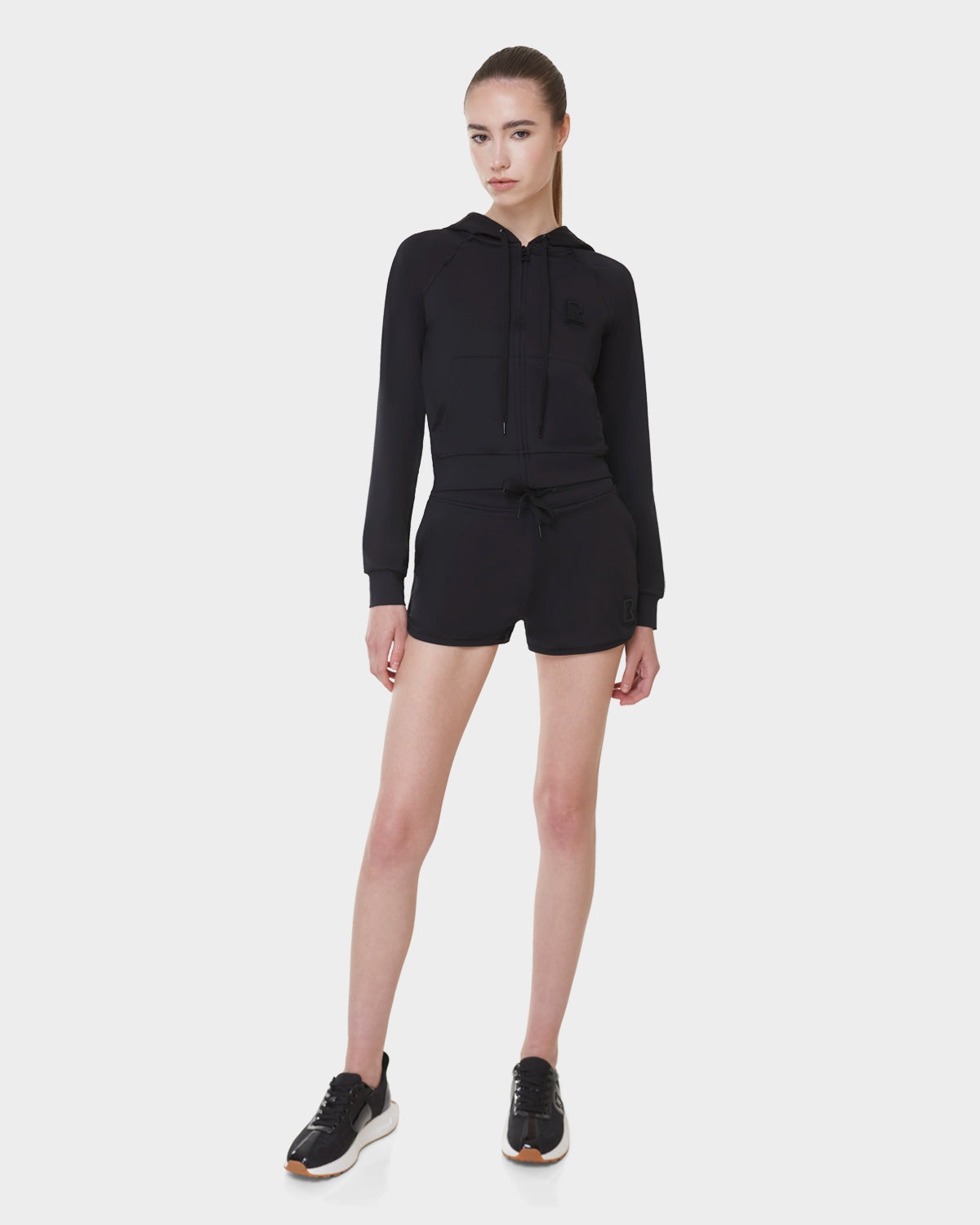 A woman with straight hair pulled back and a neutral expression stands against a white background wearing Rudsak’s SAMMY - L523014B Bottom in Black, paired with a black zip-up hoodie and black-and-white sneakers.