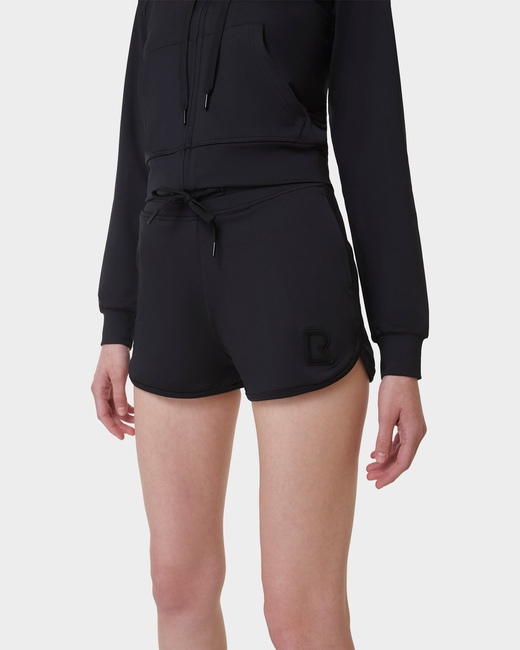 A person in a black zip-up hoodie and Rudsak SAMMY - L523014B Bottom - Black shorts with a small logo on the left leg stands against a plain white background, with their face not visible.