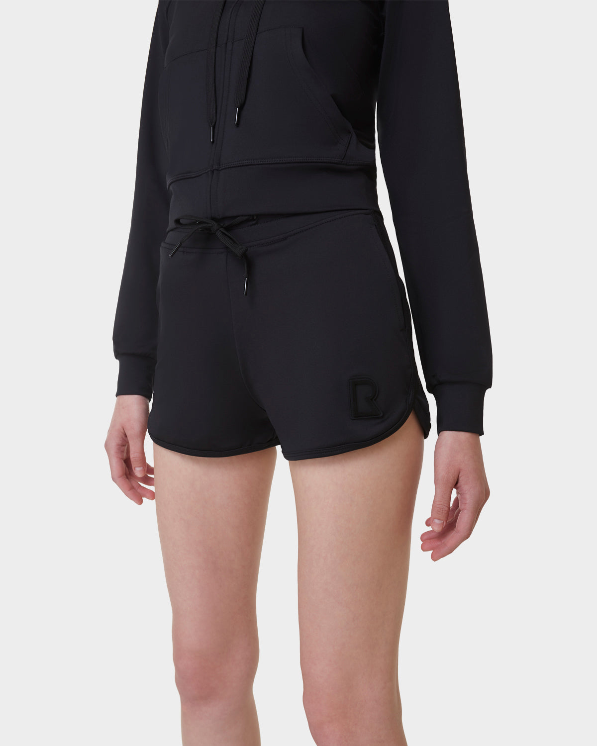 A person in a black zip-up hoodie and Rudsak SAMMY - L523014B Bottom - Black shorts with a small logo on the left leg stands against a plain white background, with their face not visible.