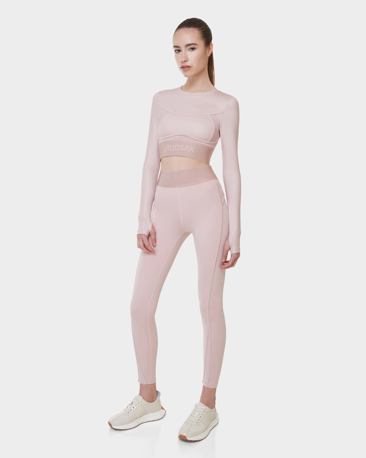 A woman faces forward in the Rudsak DOREEN - L523016T SOFT PINK long sleeve sports top and high-waisted leggings, paired with white sneakers, exuding sporty elegance against a plain white background.