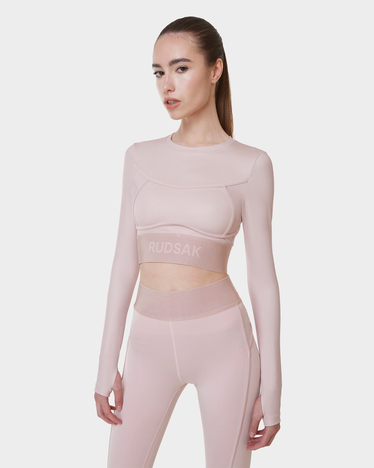 A woman models the Rudsak DOREEN - L523016T soft pink long sleeve sports top and matching high-waisted leggings with sheer panels, featuring the brand name on the crop top’s band, set against a plain white background.