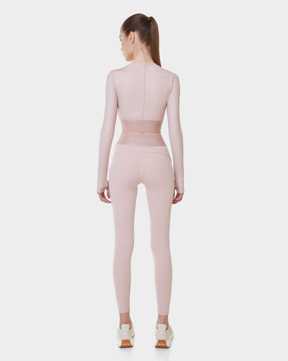 A woman faces away, wearing the Rudsak DOREEN - L523016T SOFT PINK womens long sleeve sport top and matching high-waisted leggings with RUDSAK on the waistband, her look finished with a high ponytail and white sneakers against a plain background.
