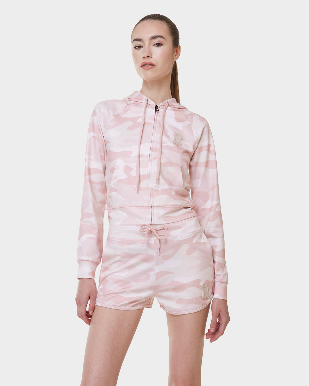 A woman wearing Rudsak's SAMMY - L523014B Camouflage Pink bottom and a matching light pink and white camo hooded jacket poses against a plain white background.