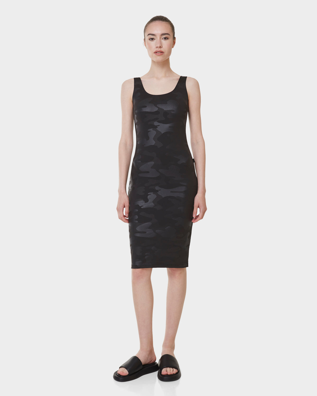 A woman faces forward in the Rudsak CLAIRE - L523026 CAMOUFLAGE - BLACK sleeveless fitted dress, knee-length with stretch fabric, paired with black slide sandals, set against a plain white background.