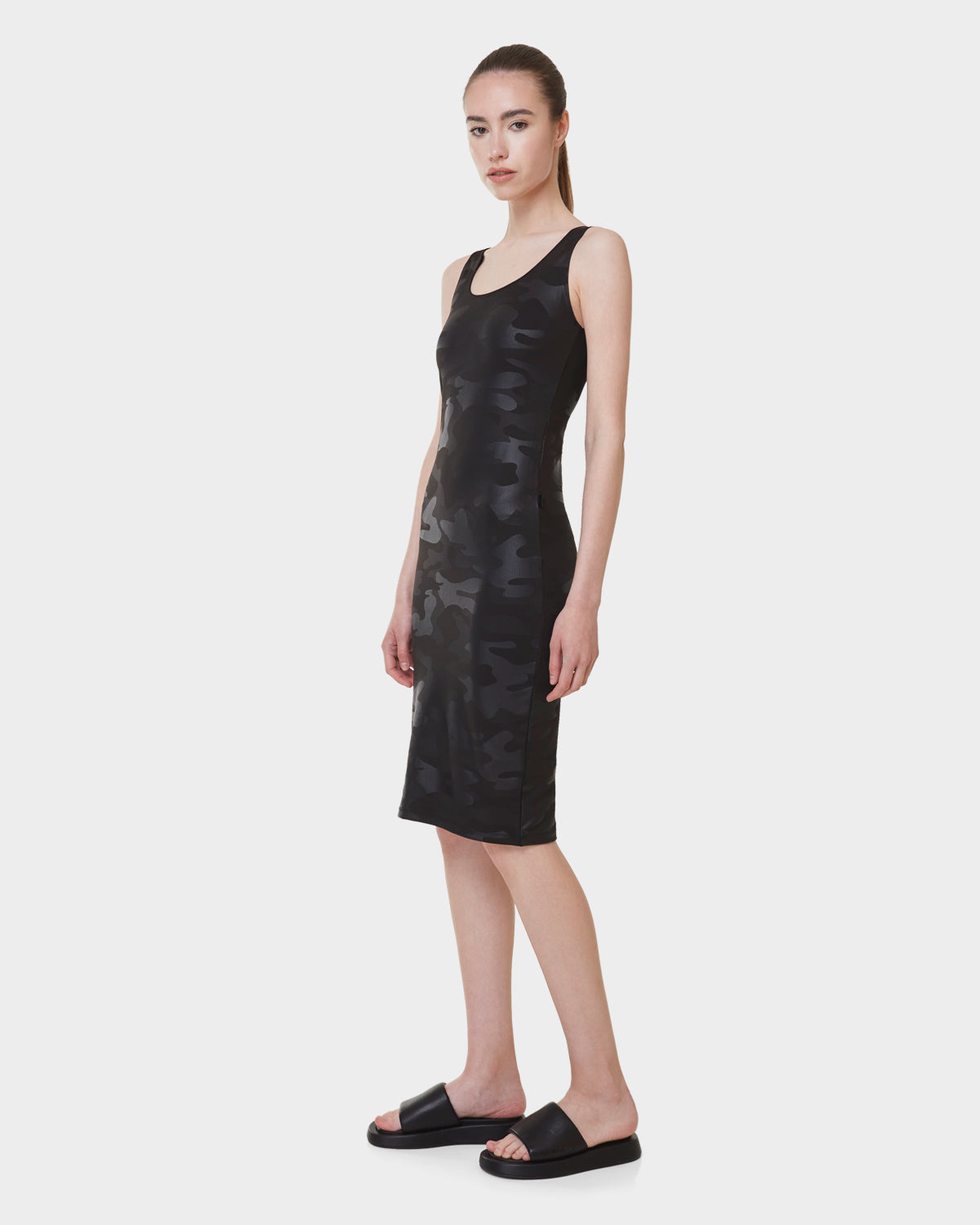 A woman stands against a white background wearing the Rudsak CLAIRE - L523026 camouflage fitted dress and black slide sandals, looking forward with a neutral expression.