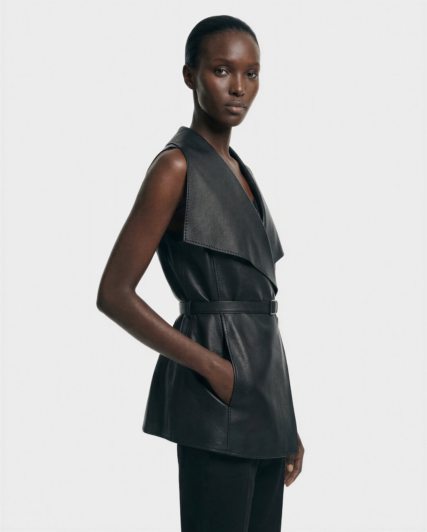 A woman poses confidently against a plain background, wearing the Rudsak ELEANOR - 8125584 Black premium full-grain leather vest with a wide collar and belted waist, paired with black pants and her hand in her pocket.