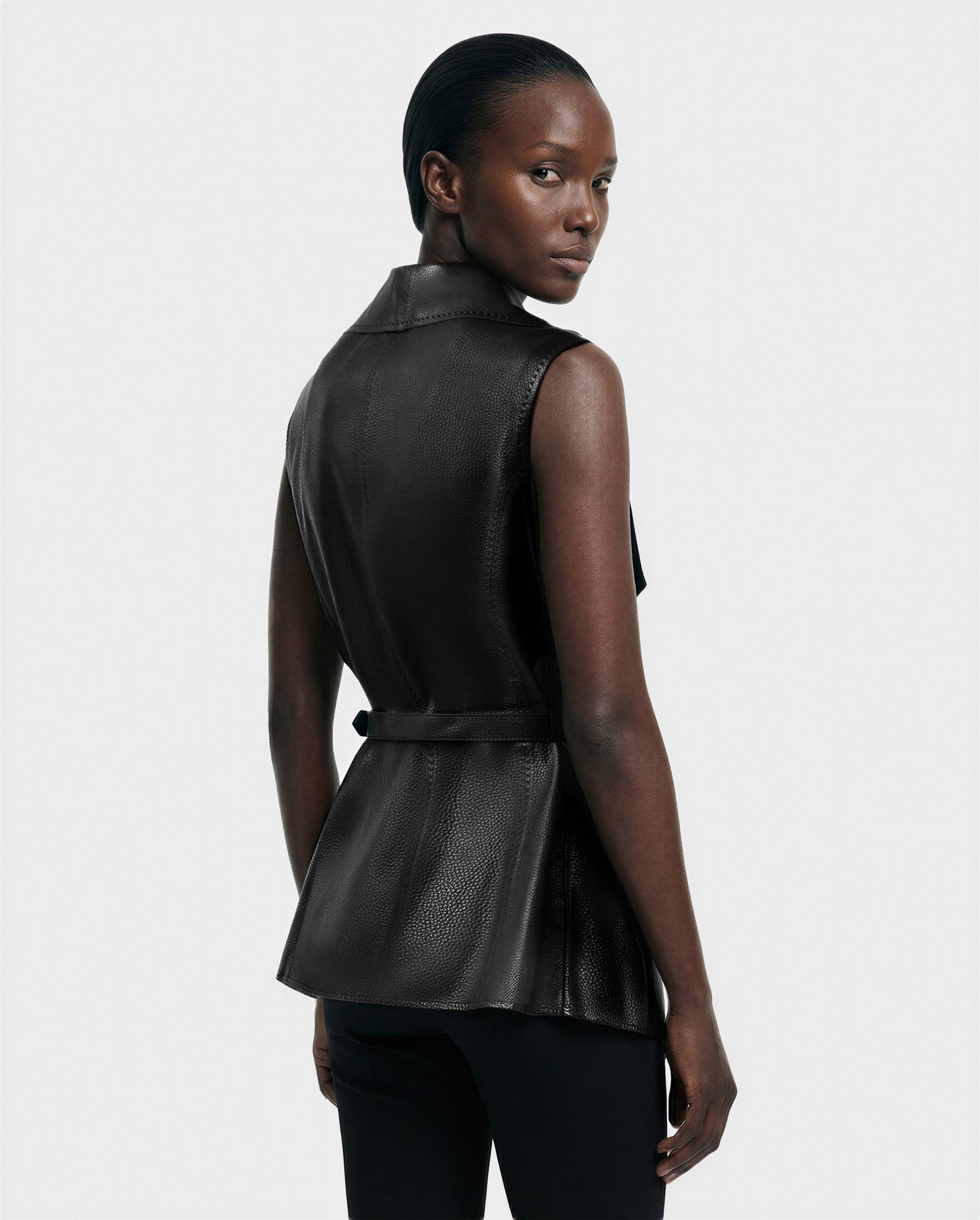 A person looks over their shoulder, wearing the Rudsak ELEANOR - 8125584 Black women's leather vest with a sleeveless cut and belted waist, paired with black pants. The background is plain white.