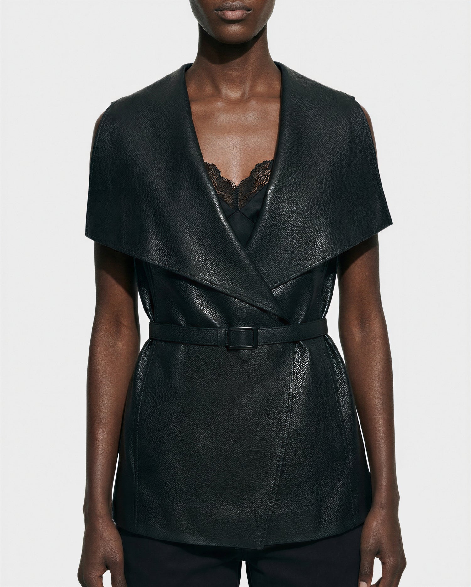 A person is wearing Rudsak’s ELEANOR - 8125584 Black belted women's leather vest with a wide collar and textured finish over a black lace-trimmed top for a sleek, modern look.