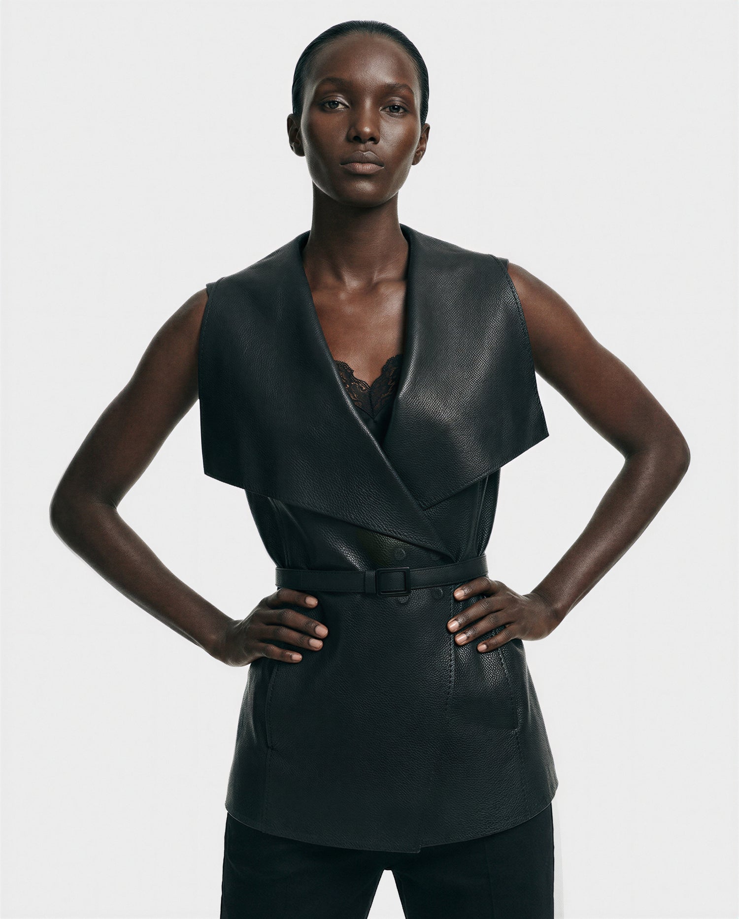 A woman stands confidently with hands on hips, wearing the Rudsak ELEANOR - 8125584 Black sleeveless full-grain leather vest with a wide collar and belted waist over a black lace top, set against a plain light background.
