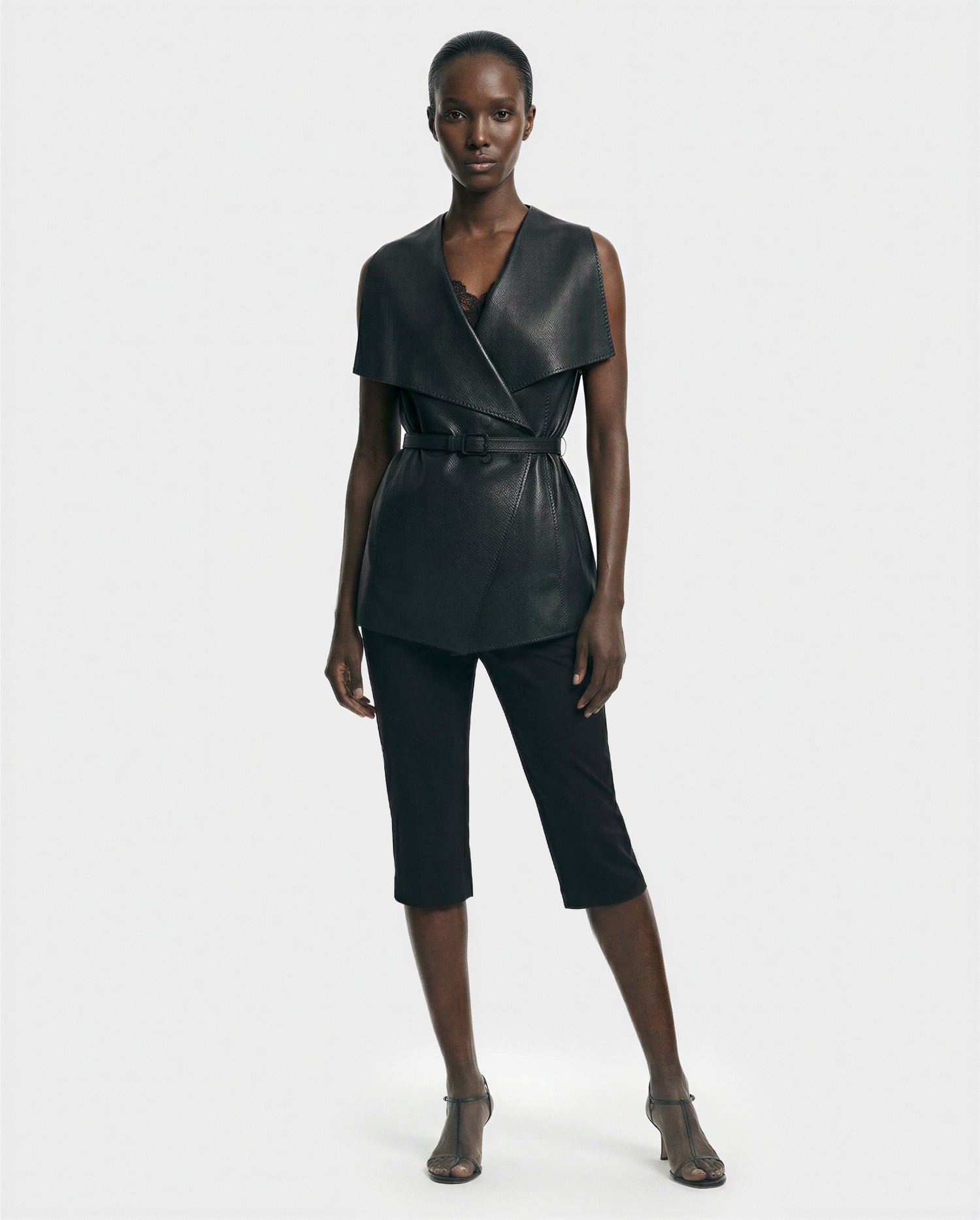 A woman stands against a plain background wearing the Rudsak ELEANOR - 8125584 Black women's leather vest, paired with black cropped pants and open-toe heels. She looks neutral, hands relaxed at her sides.