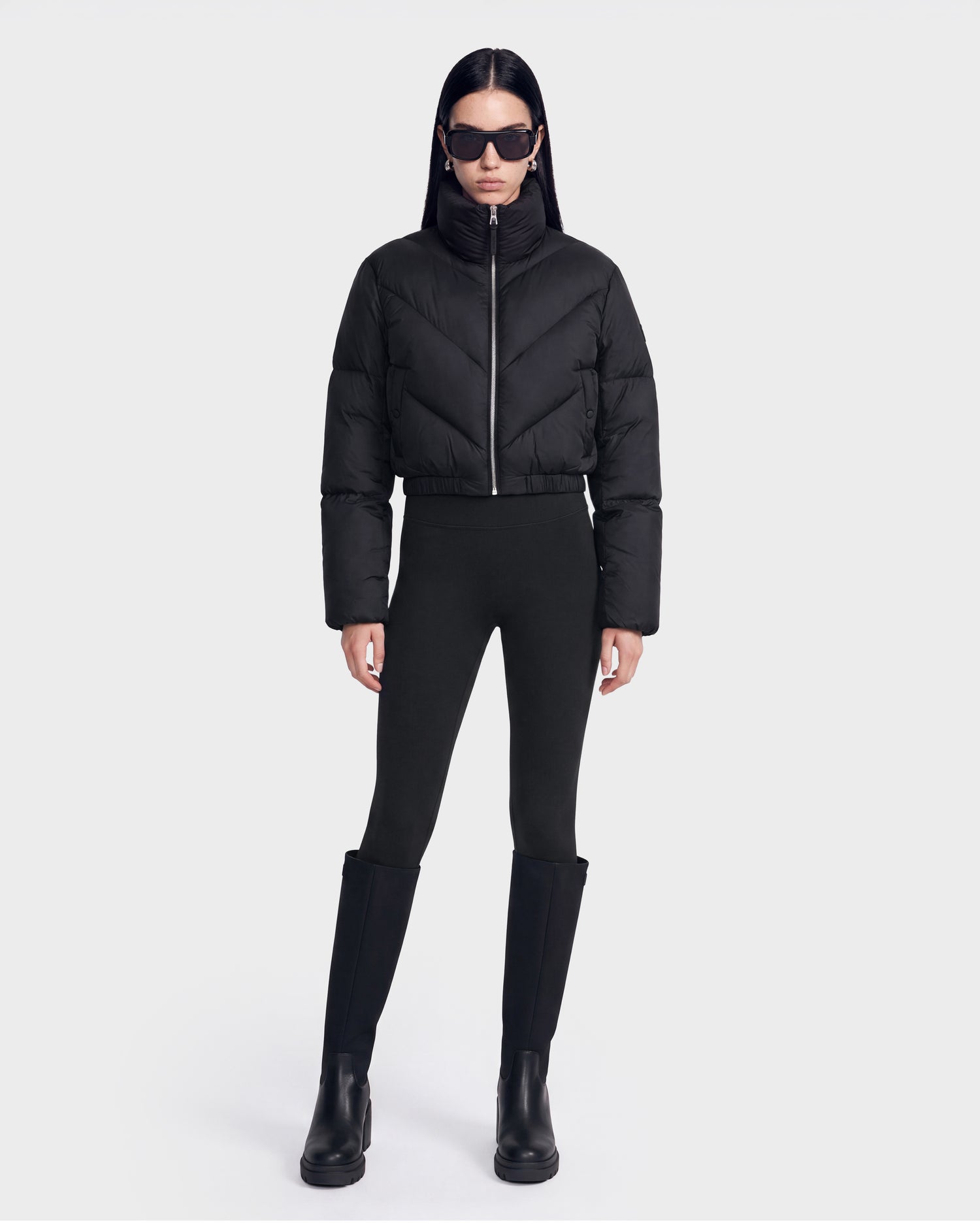 A person wearing the Rudsak ANSLIE - 8126986 Black puffer jacket, paired with black leggings, tall black boots, and sunglasses stands against a plain white background.