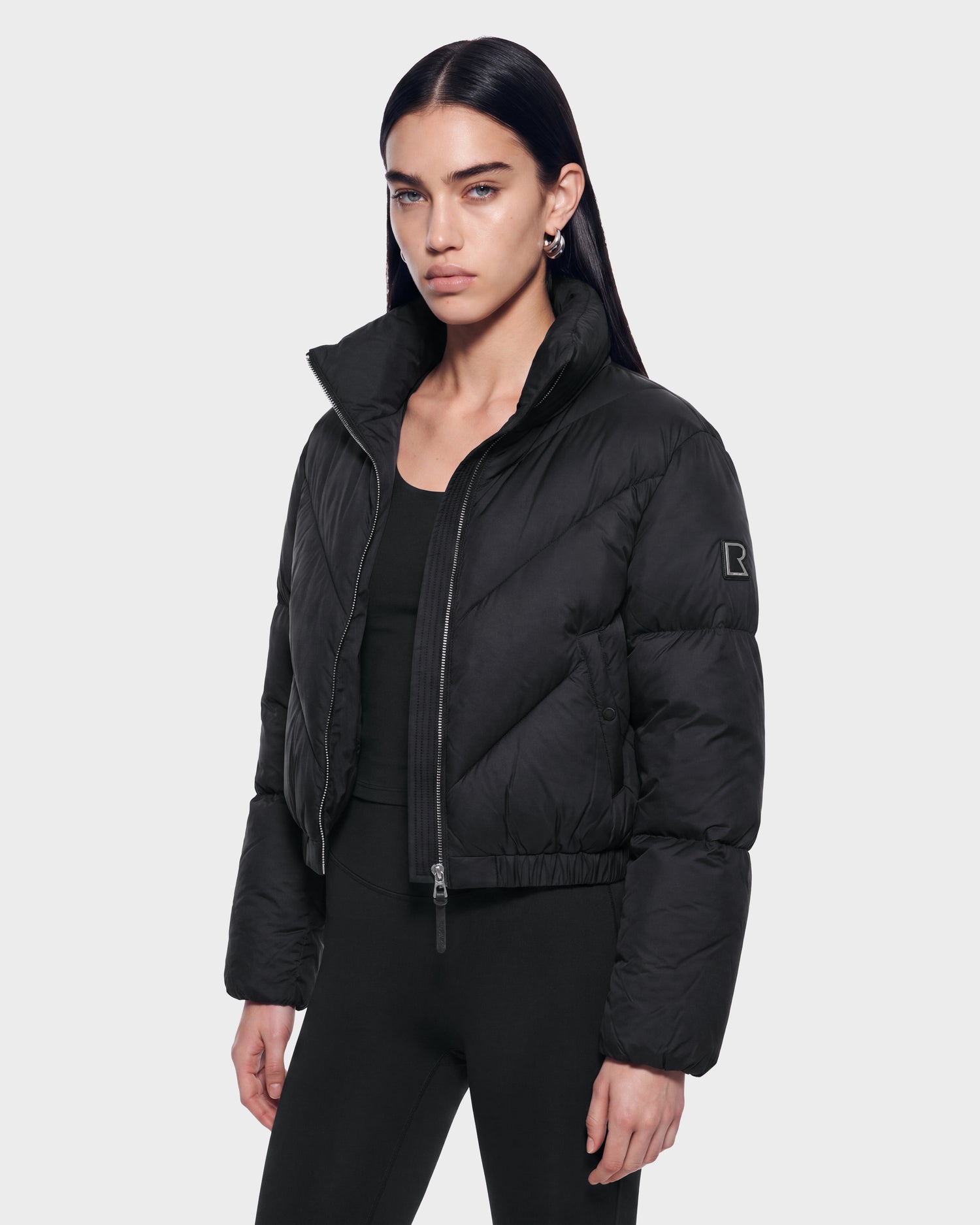 A woman with long dark hair stands against a plain light background, looking at the camera with a neutral expression. She is wearing the Rudsak ANSLIE - 8126986 Black puffer jacket and black pants.