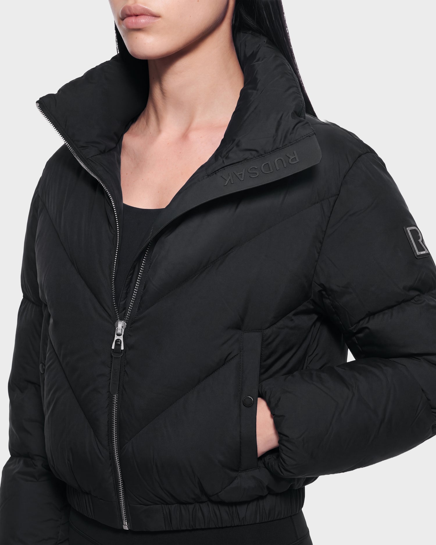 A person wearing the ANSLIE - 8126986 Black puffer jacket by Rudsak, featuring a high collar with RUDSAK branding, partially unzipped, and one hand in the pocket. The jacket also has Rudsak branding on the sleeve.