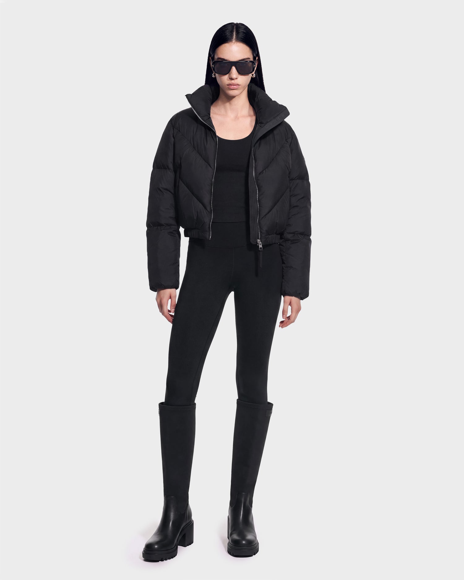 A person stands against a plain background wearing the Rudsak ANSLIE - 8126986 Black puffer jacket, paired with a black top, black leggings, knee-high black boots, and dark sunglasses.