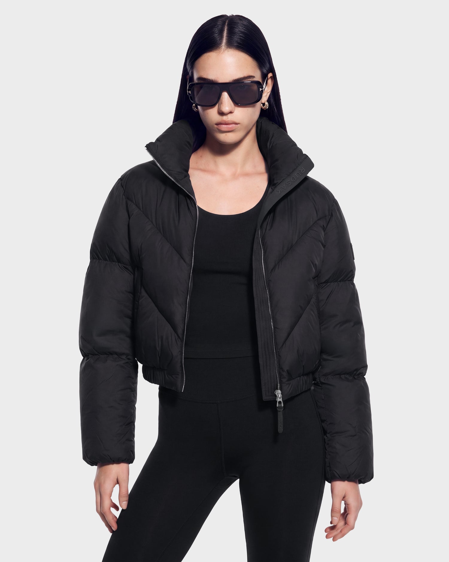 A woman stands against a plain light background wearing the ANSLIE - 8126986 Black puffer jacket by Rudsak, paired with a black top and black leggings. She accessorizes her look with large black sunglasses and small hoop earrings.