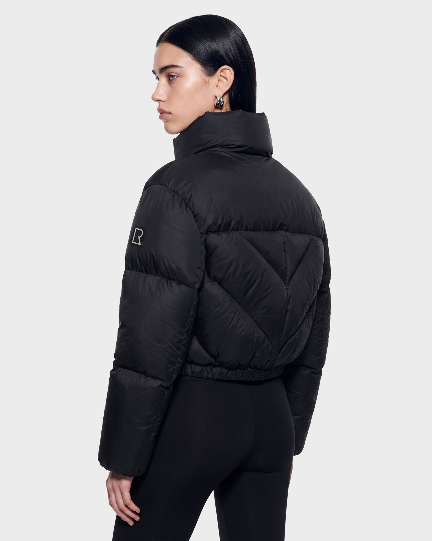 A woman with straight dark hair models the Rudsak ANSLIE - 8126986 Black puffer jacket, featuring a high collar and fitted black pants, as she stands against a plain light background, facing away while turning her head to the side.