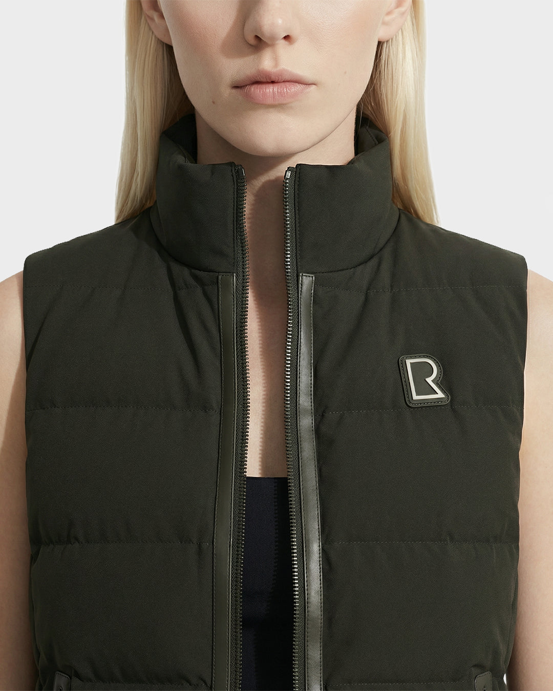 A woman wears the Rudsak GABRIELA - 8125014 dark olive sleeveless lightweight puffer vest with a high collar and R logo on the chest; the unzipped women’s down vest reveals a black top underneath.