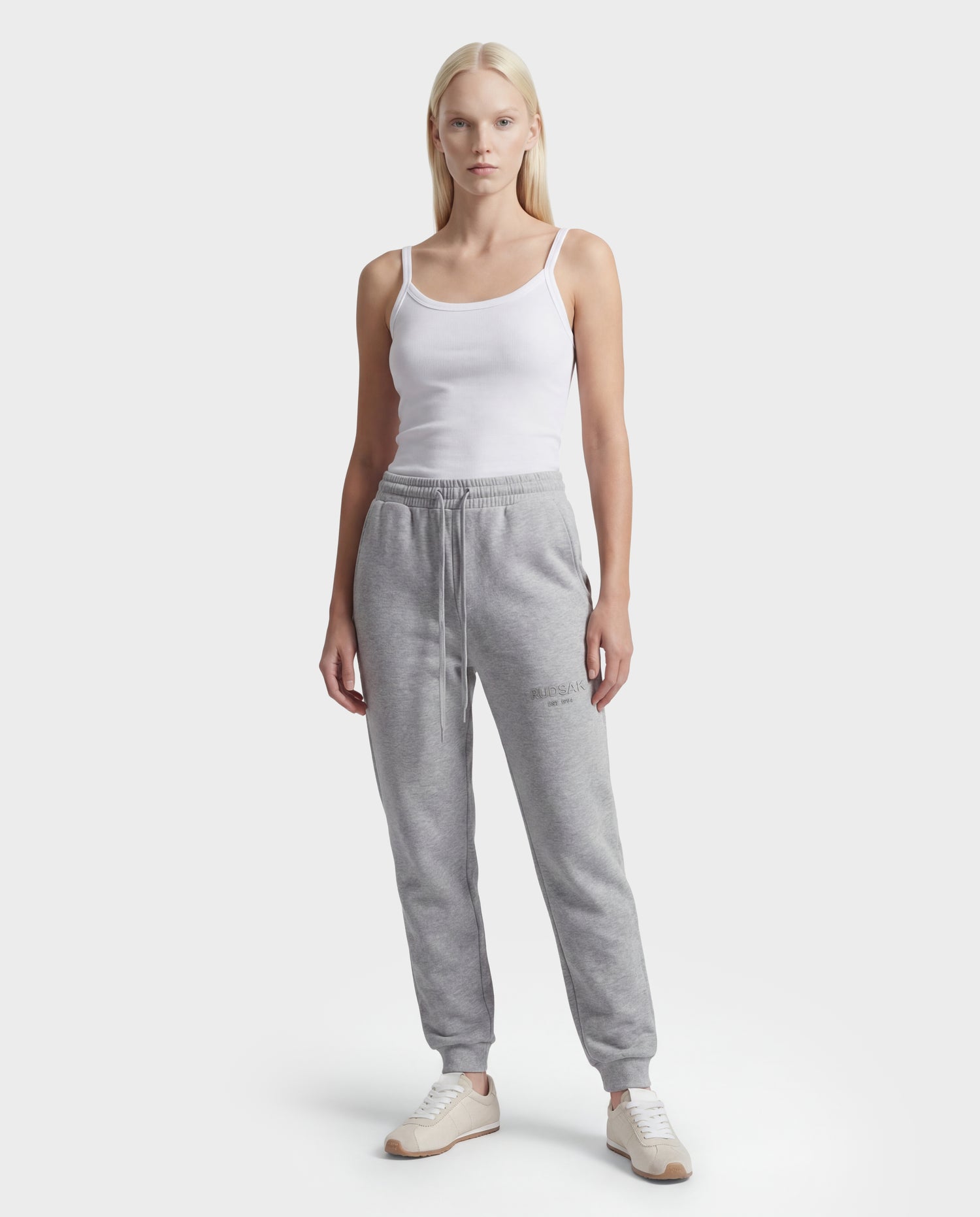 A woman stands facing forward in a white tank top, beige sneakers, and Rudsak's JOSS - 9524009 Heather Grey W stylish joggers against a plain light background.