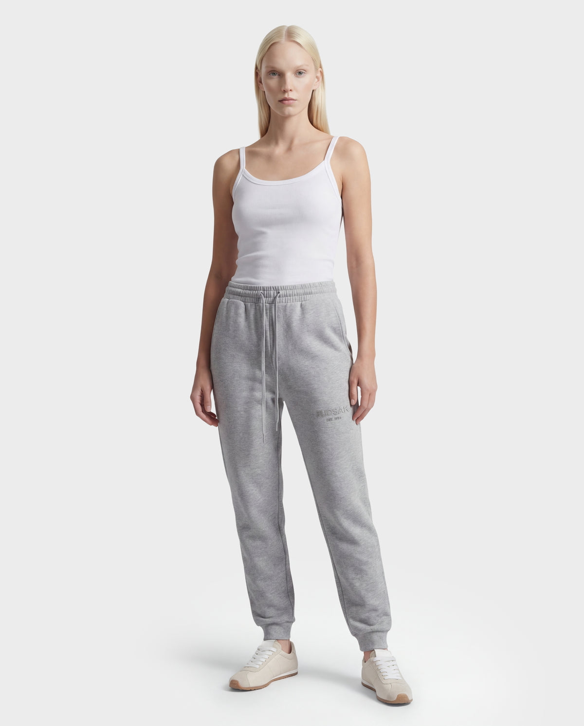 A woman stands facing forward in a white tank top, beige sneakers, and Rudsak's JOSS - 9524009 Heather Grey W stylish joggers against a plain light background.