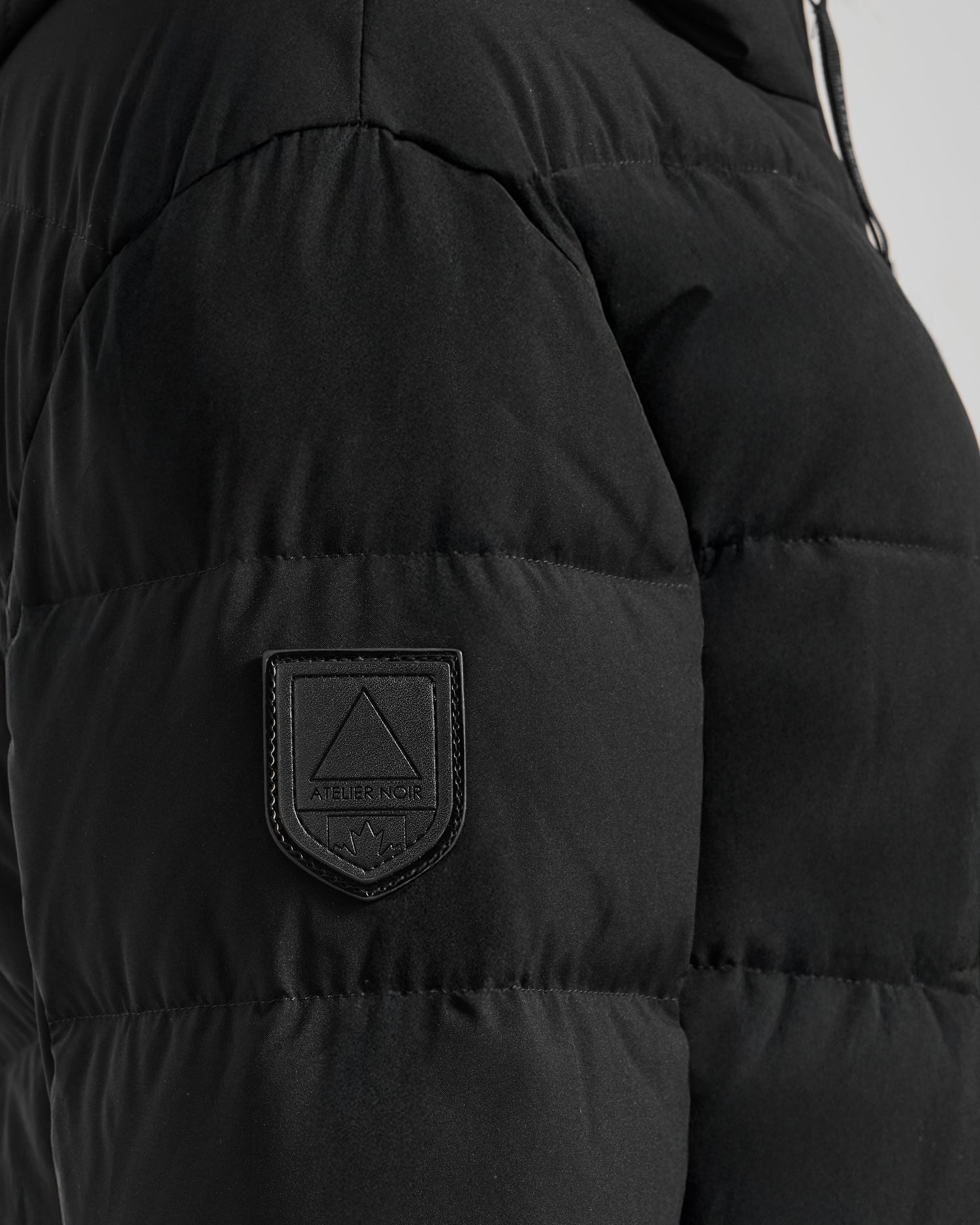 Close-up of the Rudsak LAUREN - 4124520 Black puffer jacket, featuring quilted matte water-repellent fabric and a stitched sleeve patch with a triangle logo and "Atelier Noir" beneath it.