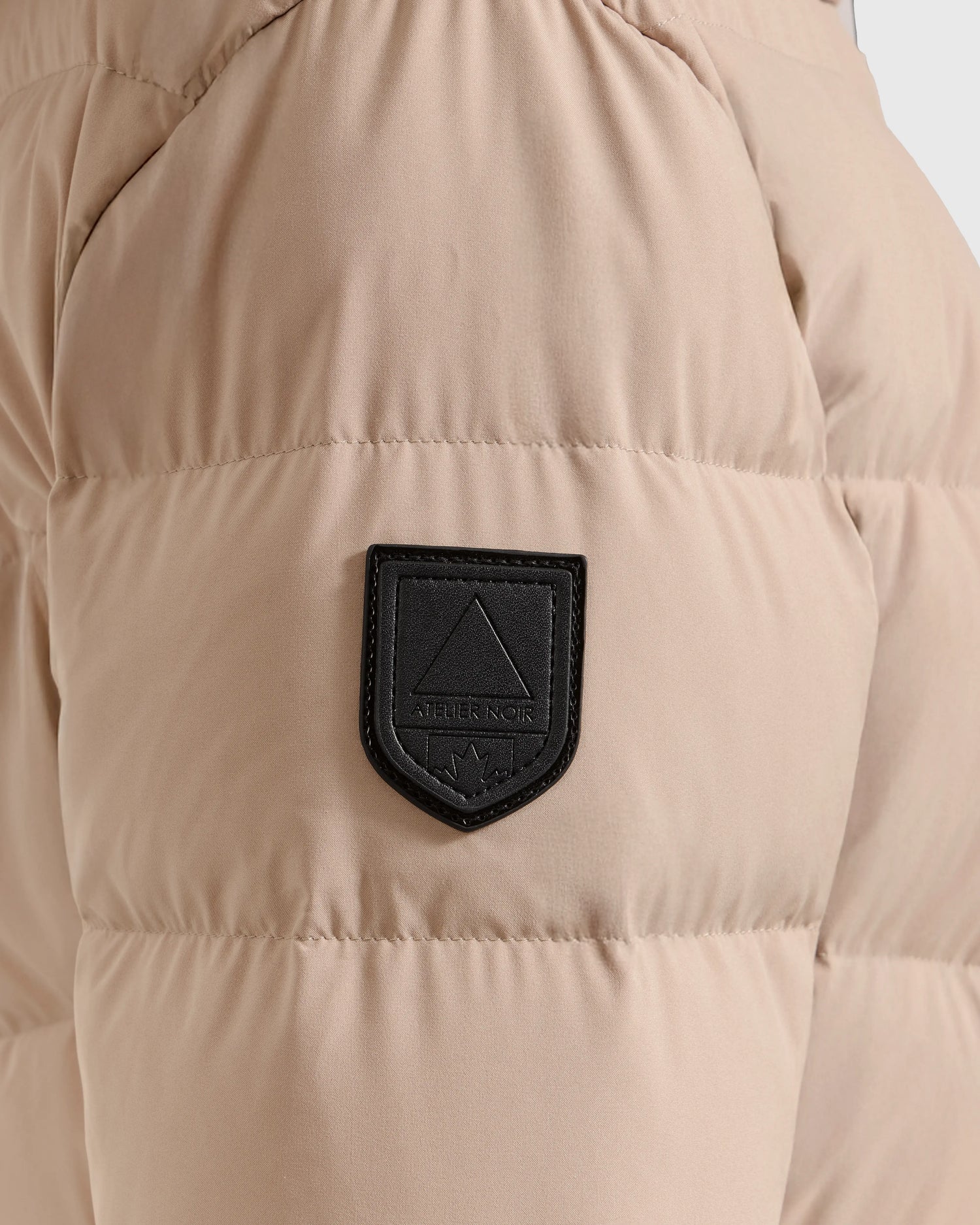 Close-up of the Rudsak LAUREN - 4124520 Taupe puffer jacket, featuring beige water-repellent quilted fabric and a branded black sleeve patch. This padded coat is designed to keep you warm on cold days.