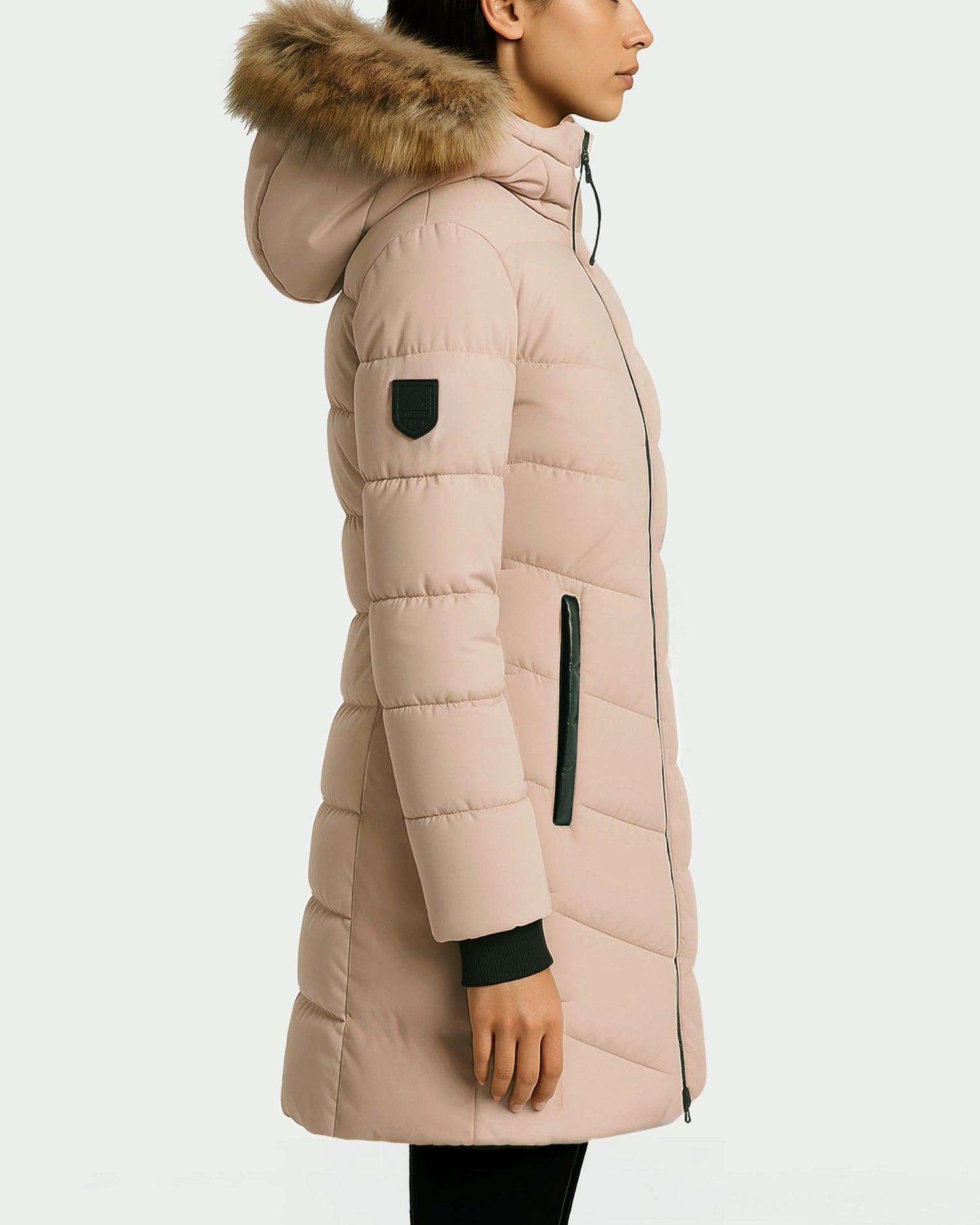 A woman stands in profile wearing the Rudsak LAUREN - 4124520 Taupe, a knee-length, water-repellent puffer coat with faux fur hood, black zipper details, and a black patch on the upper sleeve against a plain background.